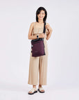 Full body view of a dark haired model facing the camera and smiling. She is wearing a tan pinstriped jumpsuit, white sandals and Sherpani's Anti-Theft bag, the Geo AT in Merlot, as a crossbody.