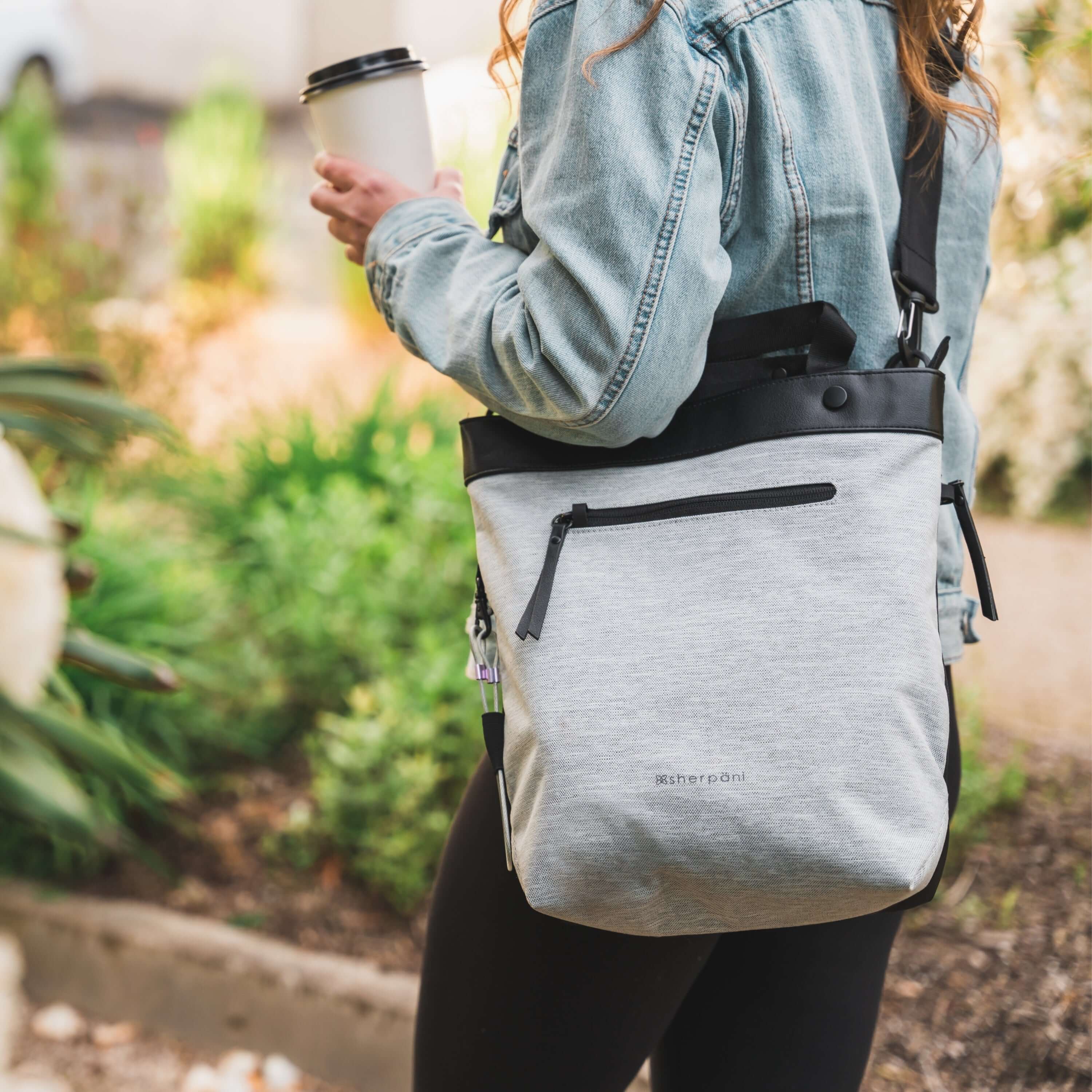 Close up view of a blonde haired model standing outside holding a coffee. She is wearing a jean jacket, black leggings and Sherpani&#39;s Anti-Theft bag, the Geo AT in Sterling, as a crossbody.