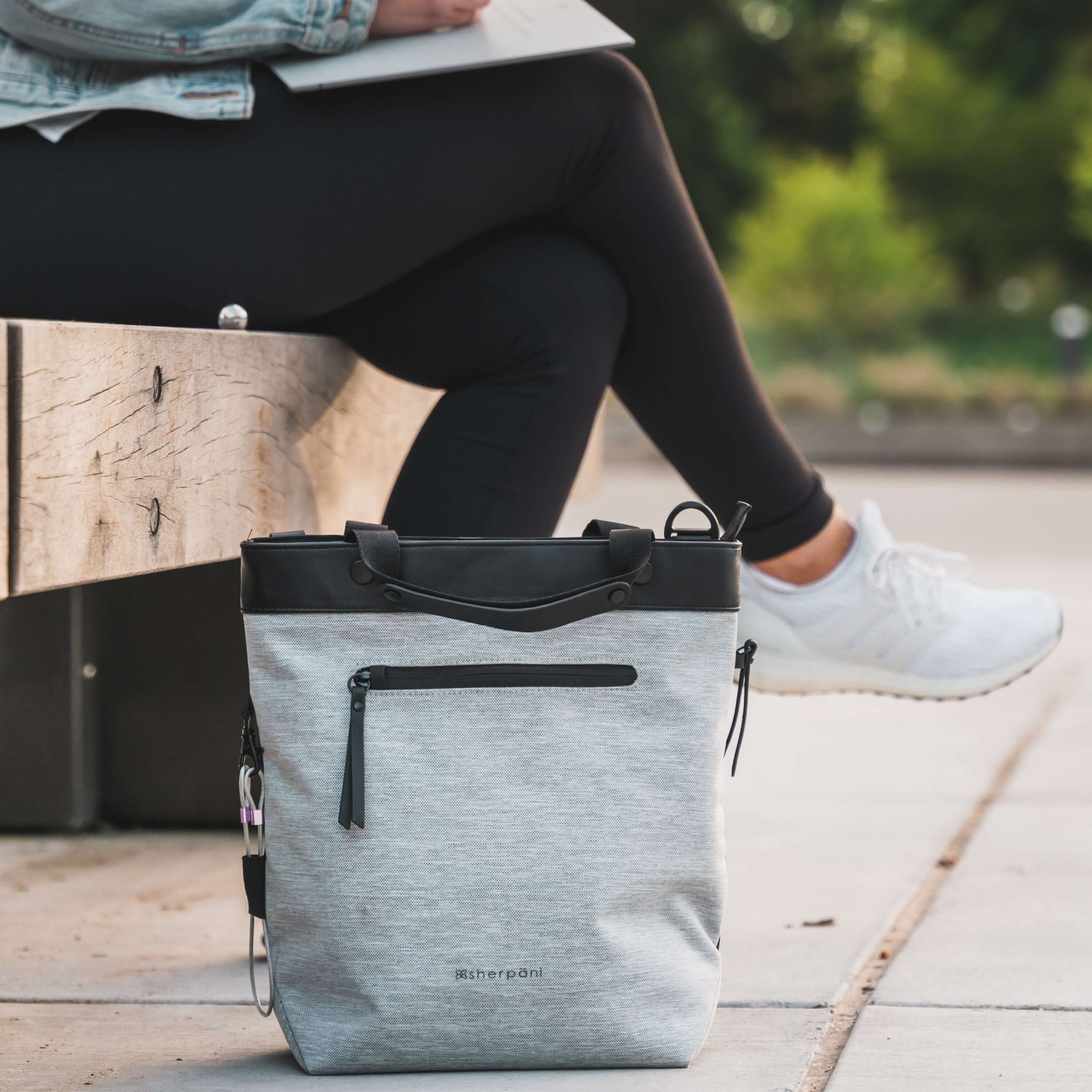 Close up view of a model&#39;s lower body sitting on a bench outside. She is wearing black leggings and white shoes. On the ground in front of her sits Sherpani&#39;s Anti-Theft bag the Geo AT in Sterling.