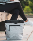 Close up view of a model's lower body sitting on a bench outside. She is wearing black leggings and white shoes. On the ground in front of her sits Sherpani's Anti-Theft bag the Geo AT in Sterling.