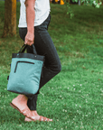 Close up view of a model's lower body. She is facing the side and walking on the grass. She wears a white shirt, black pants and tan sandals. She is carrying Sherpani's Ant-Theft bag, the Geo AT in Teal, by the tote handles.