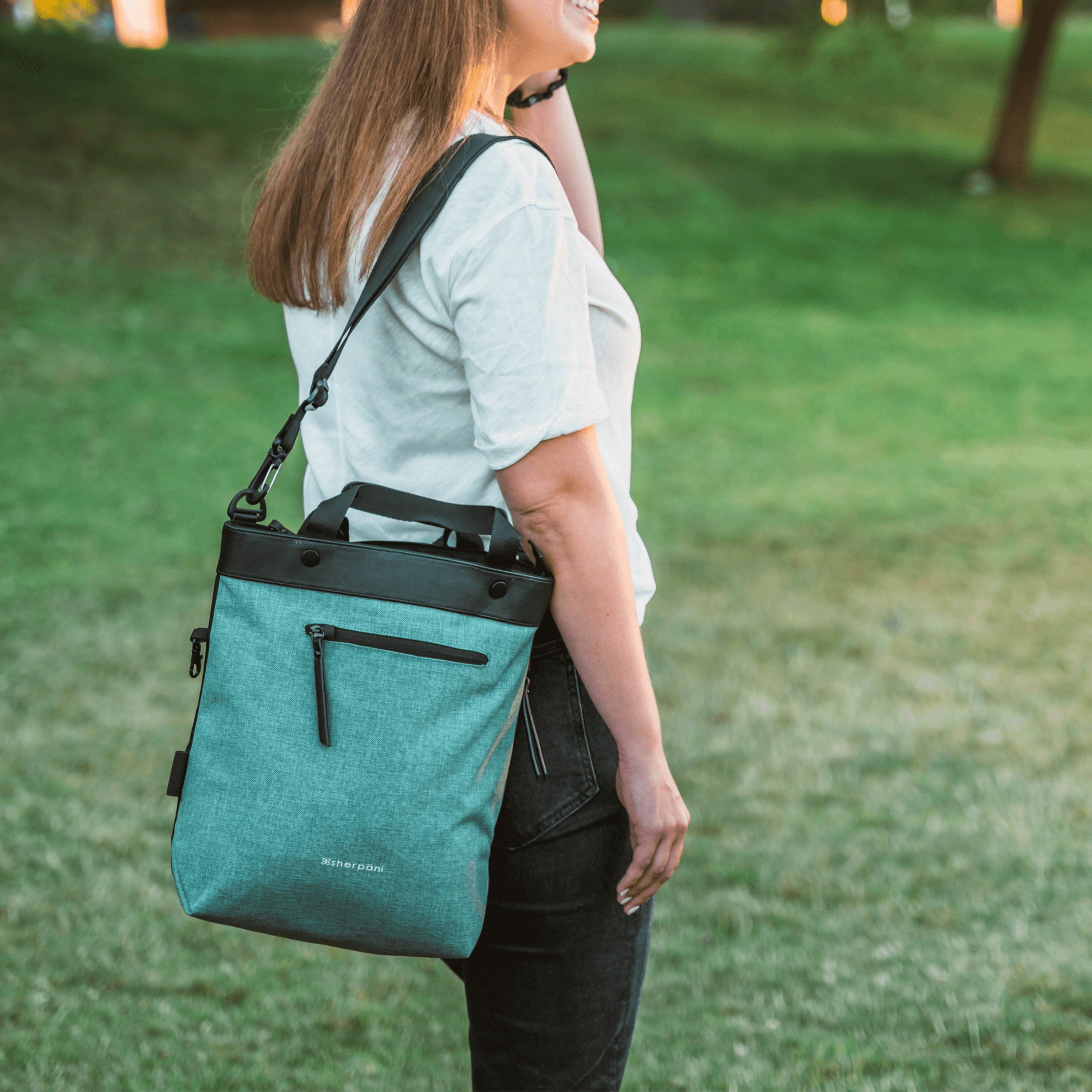 A blonde haired model is standing outside in a park, facing to the side and smiling. She is wearing a white tee shirt, black jeans and has Sherpani&#39;s Anti-Theft bag, the Geo AT in Teal, over her shoulder.