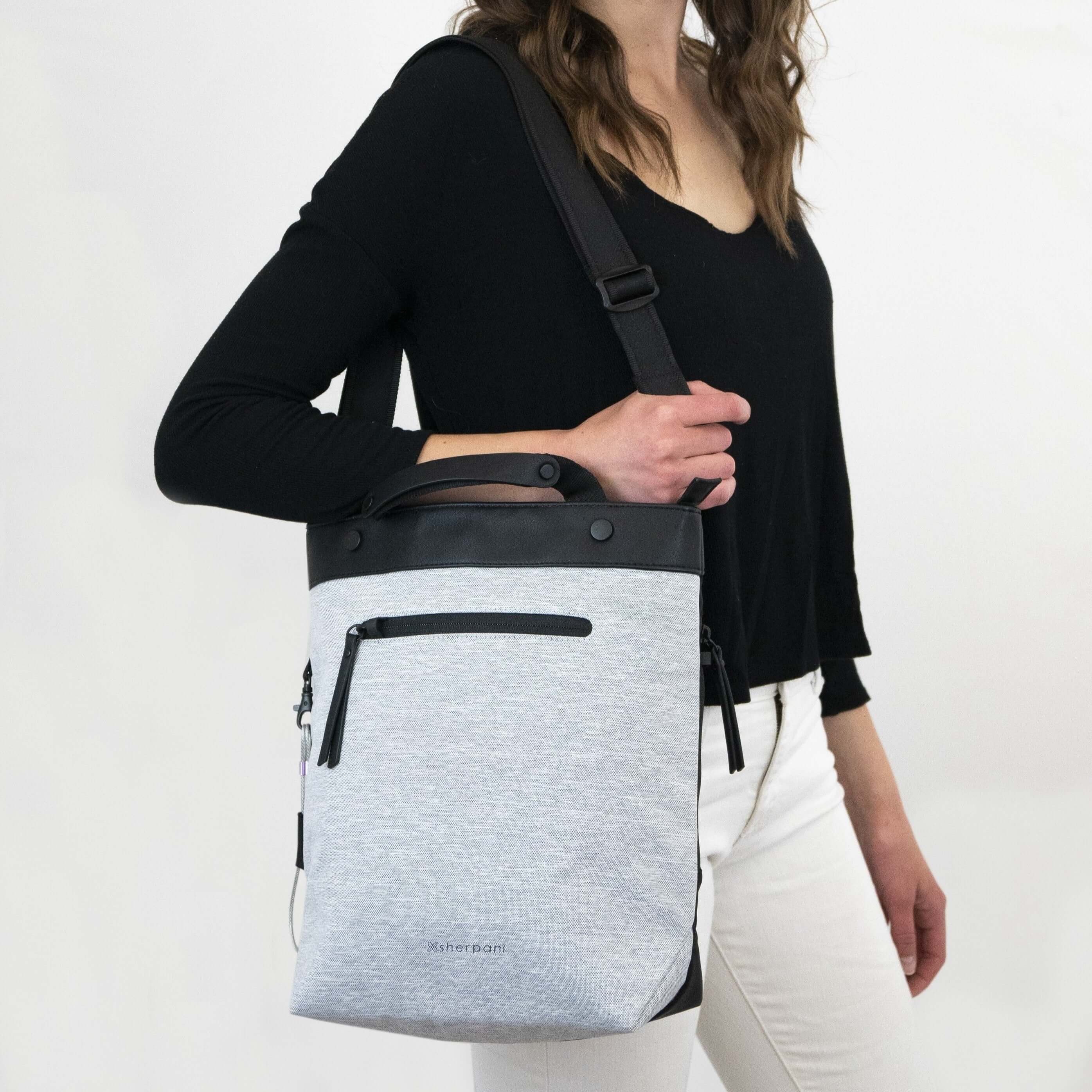 Close up view of a brown haired model wearing a black shirt, white pants and Sherpani&#39;s Anti-Theft bag, the Geo AT in Sterling, over her shoulder.