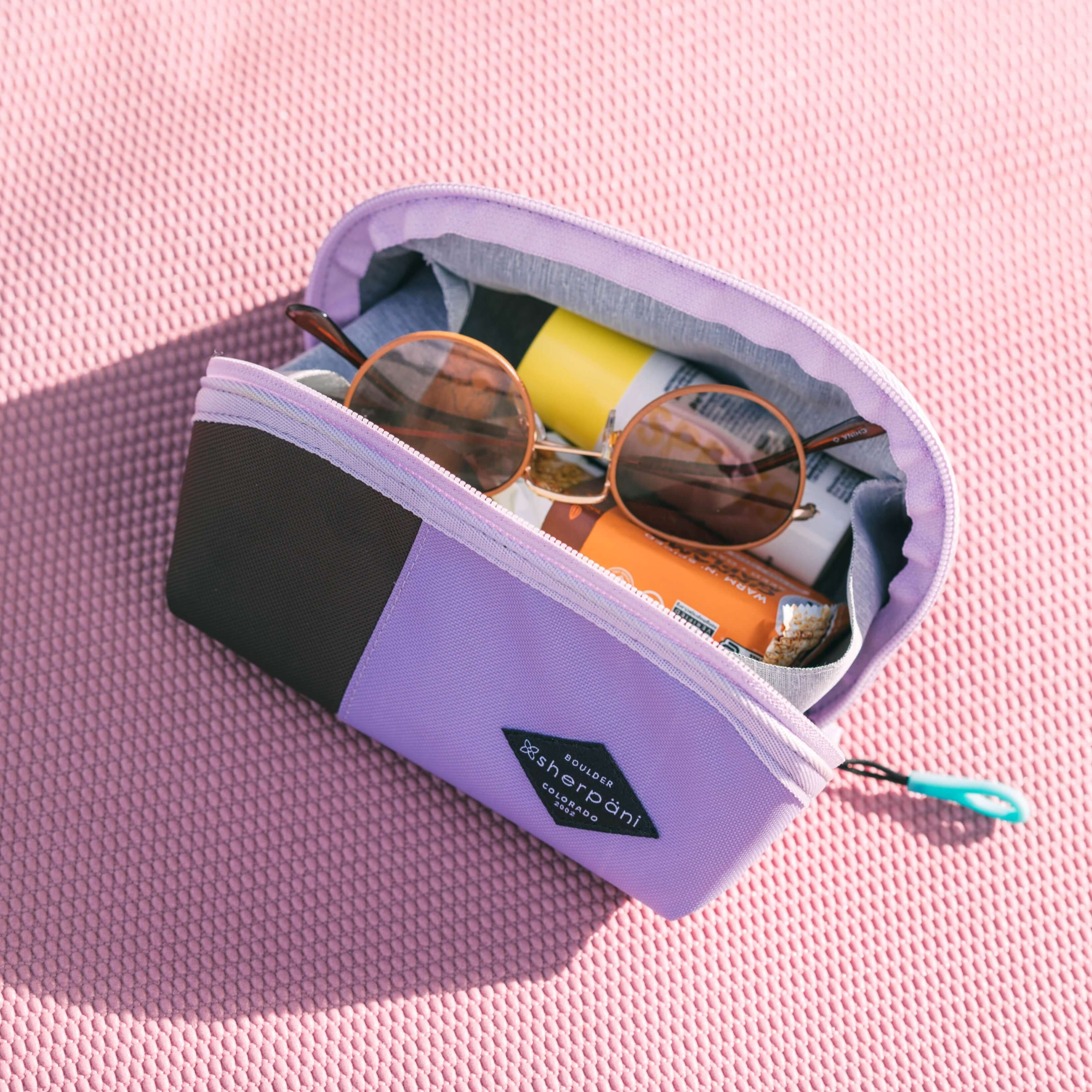 Top view of Sherpani travel accessory, the Harmony in Lavender. The pouch is open to reveal several items: sunglasses, granola bar and sunscreen. 