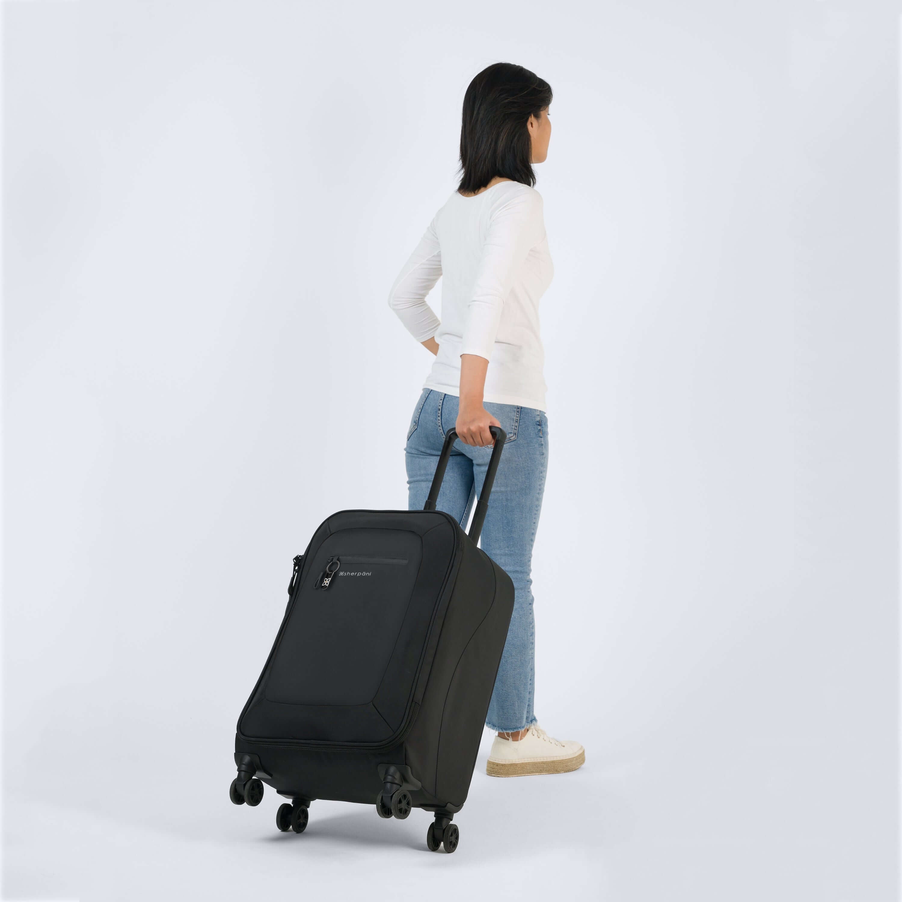 Full body view of a dark haired model facing away from the camera. She is wearing a white shirt, jeans, and white shoes. She is pulling Sherpani's Anti-Theft luggage, the Hemisphere in Carbon, along behind her. 
