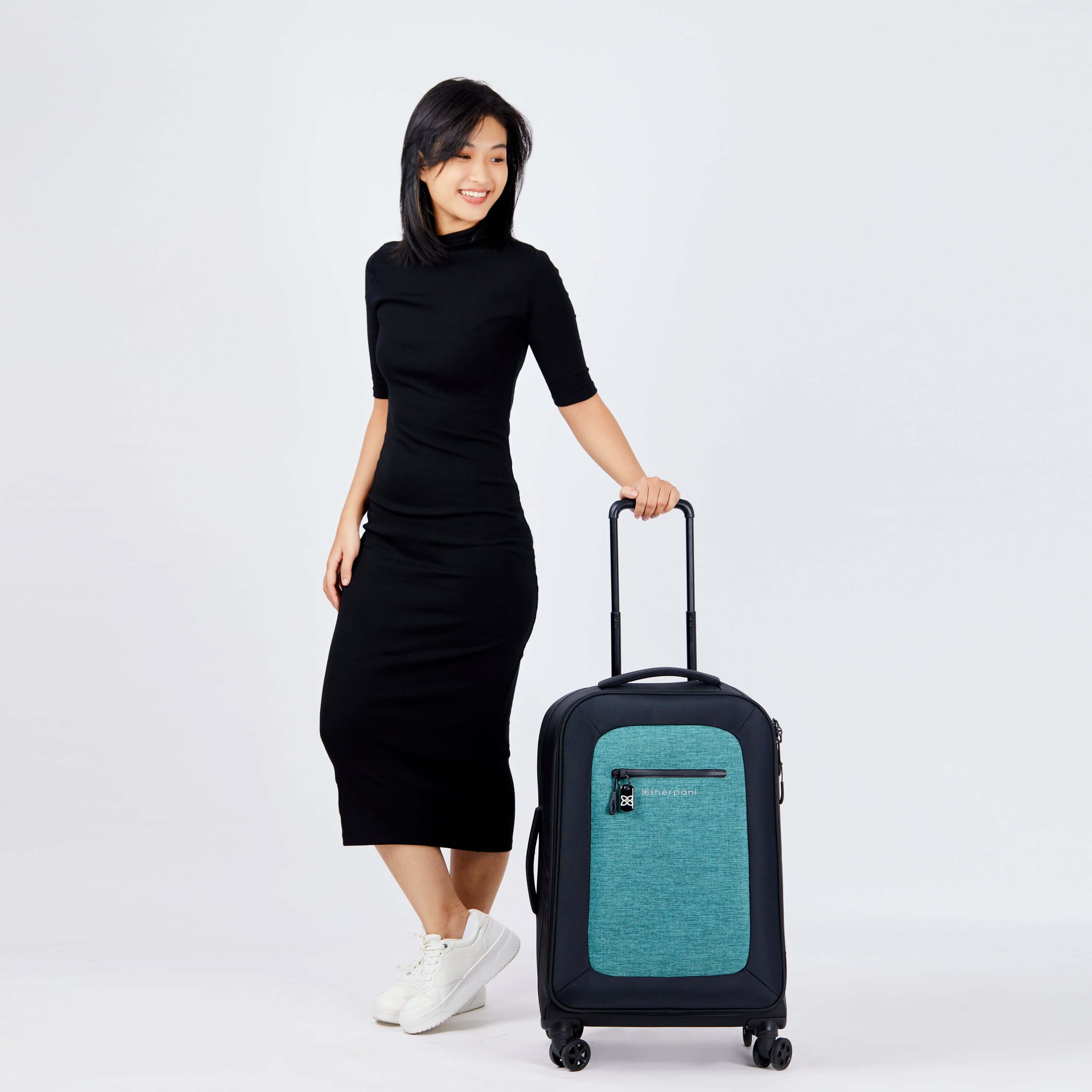 Full body view of a dark haired model smiling over her left shoulder. She wears a black dress and white sneakers. She is holding the retractable handle of Sherpani's Anti-Theft luggage, the Hemisphere in Teal, which stands next to her. 