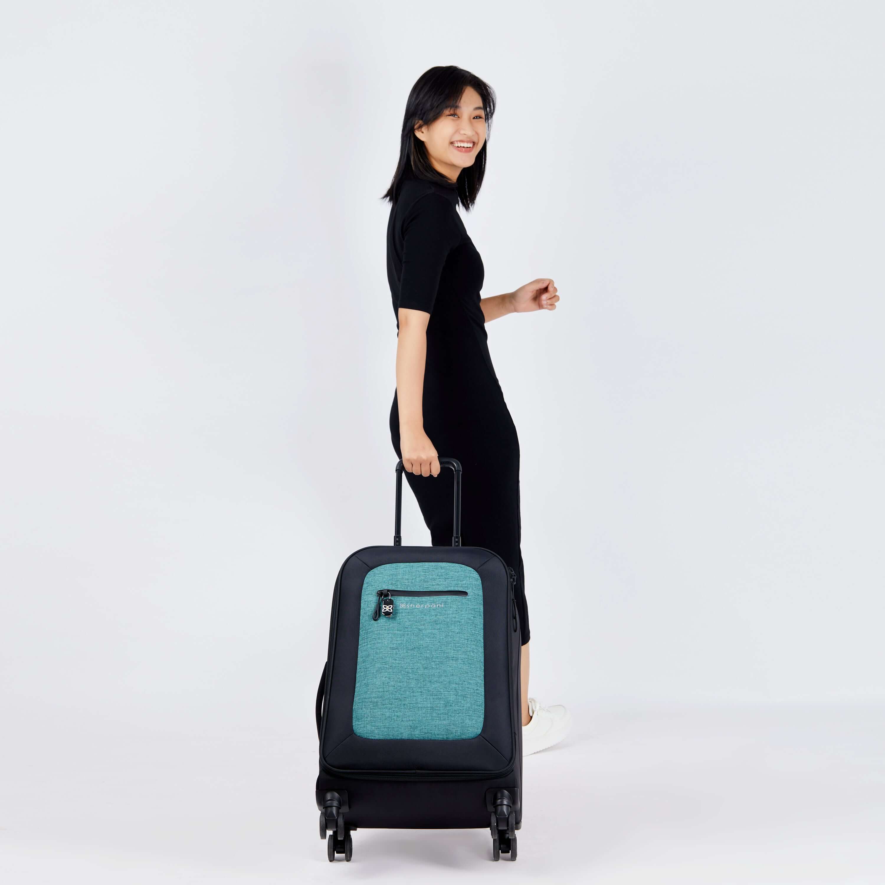 Full body view of a dark haired model facing away from the camera and smiling over her right shoulder. She is wearing a black dress and white sneakers. She is pulling Sherpani's Anti-Theft luggage, the Hemisphere in Teal, along behind her. 