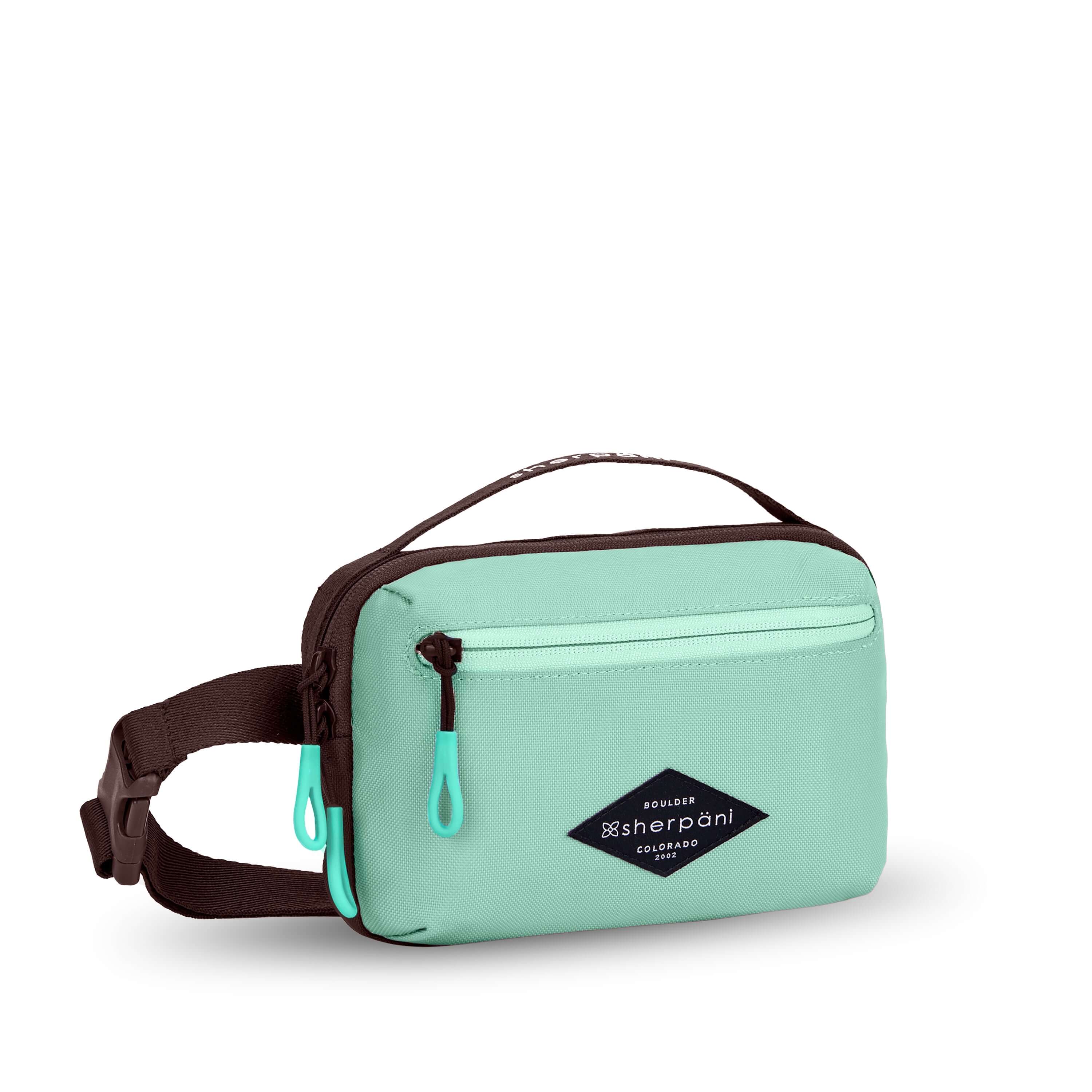 Angled front view of Sherpani’s fanny pack, the Hyk in Seagreen. The bag is two-toned, the front half is light green and the back half is brown. There is an external zipper pocket on the front of the bag. Easy-pull zippers are accented in light green. The fanny pack features an adjustable strap with a buckle.