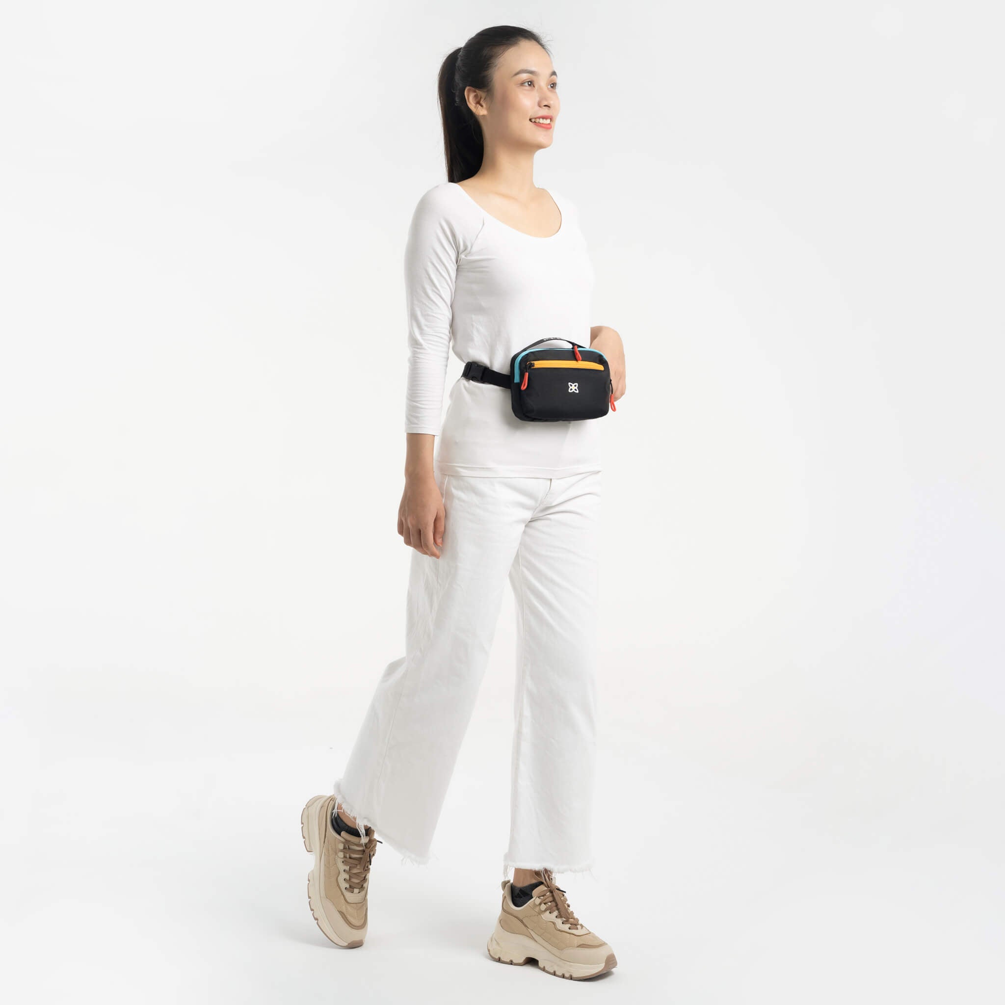 Full body view of a dark haired model smiling. She is wearing a white tee shirt, white pants, and Sherpani&#39;s fanny pack, the Hyk in Chromatic, as a fanny bag..
