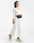 Full body view of a dark haired model smiling. She is wearing a white tee shirt, white pants, and Sherpani's fanny pack, the Hyk in Chromatic, as a fanny bag..