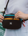 Close up view of a woman's hands opening the zipper pocket of Sherpani waist pack, the Hyk in Chromatic.