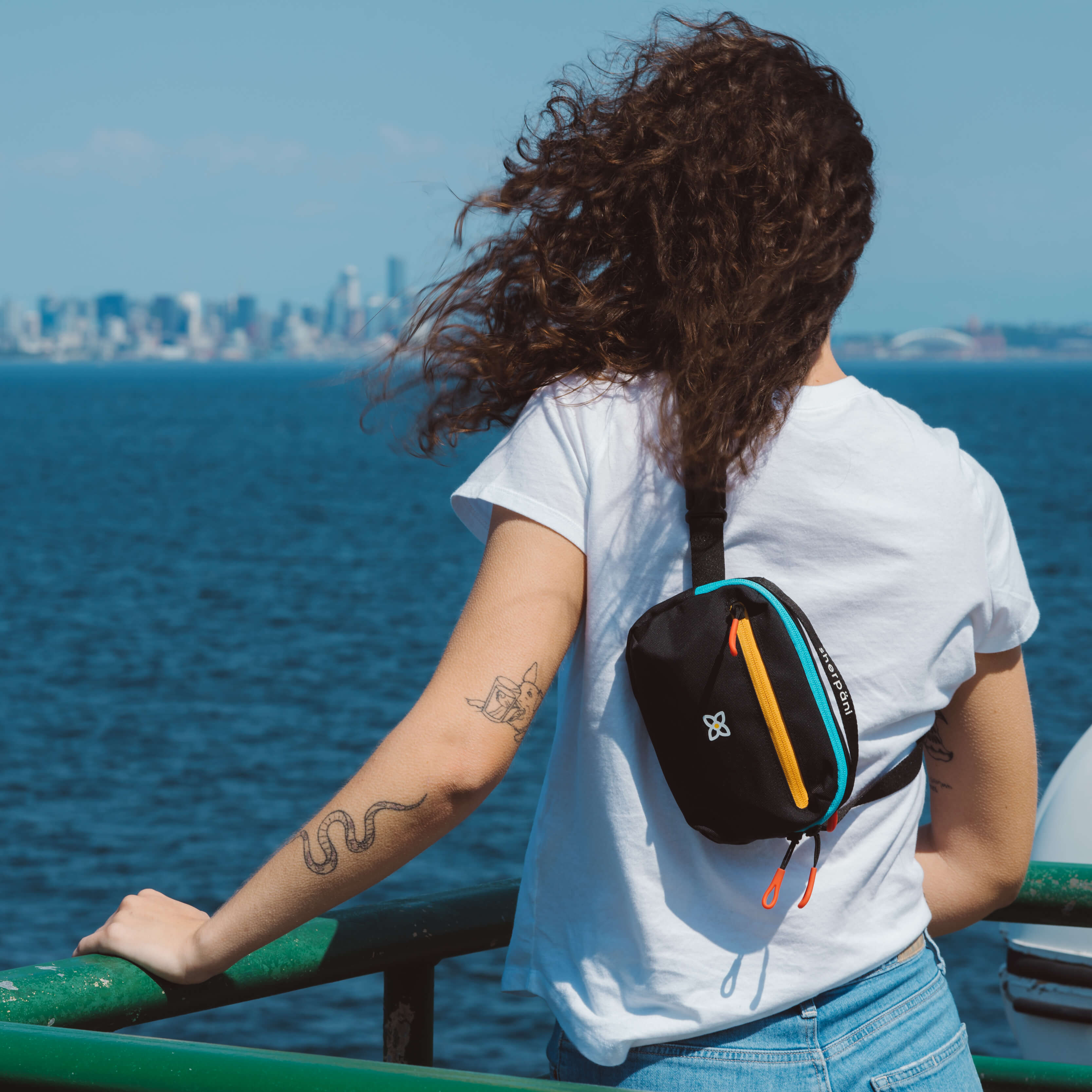 A traveling woman stands in front of a waterfront wearing Sherpani hip pack, the Hyk in Chromatic, as a mini crossbody travel purse.