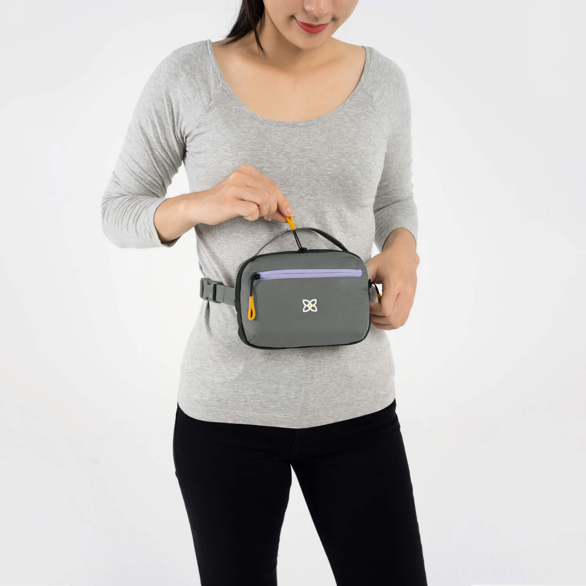 A model is opening the zipper pocket of Sherpani belt pack, the Hyk in Moonstone, which she wears as a small fanny pack. 