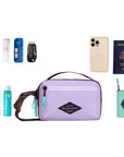 Top view of example items to fill the bag. Sherpani's fanny pack, the Hyk in Lavender, lies in the center. It is surrounded by an assortment of items: skincare products, sunscreen, beauty spray, mints, car key, phone, passport and Sherpani travel accessory the Barcelona in Seagreen.