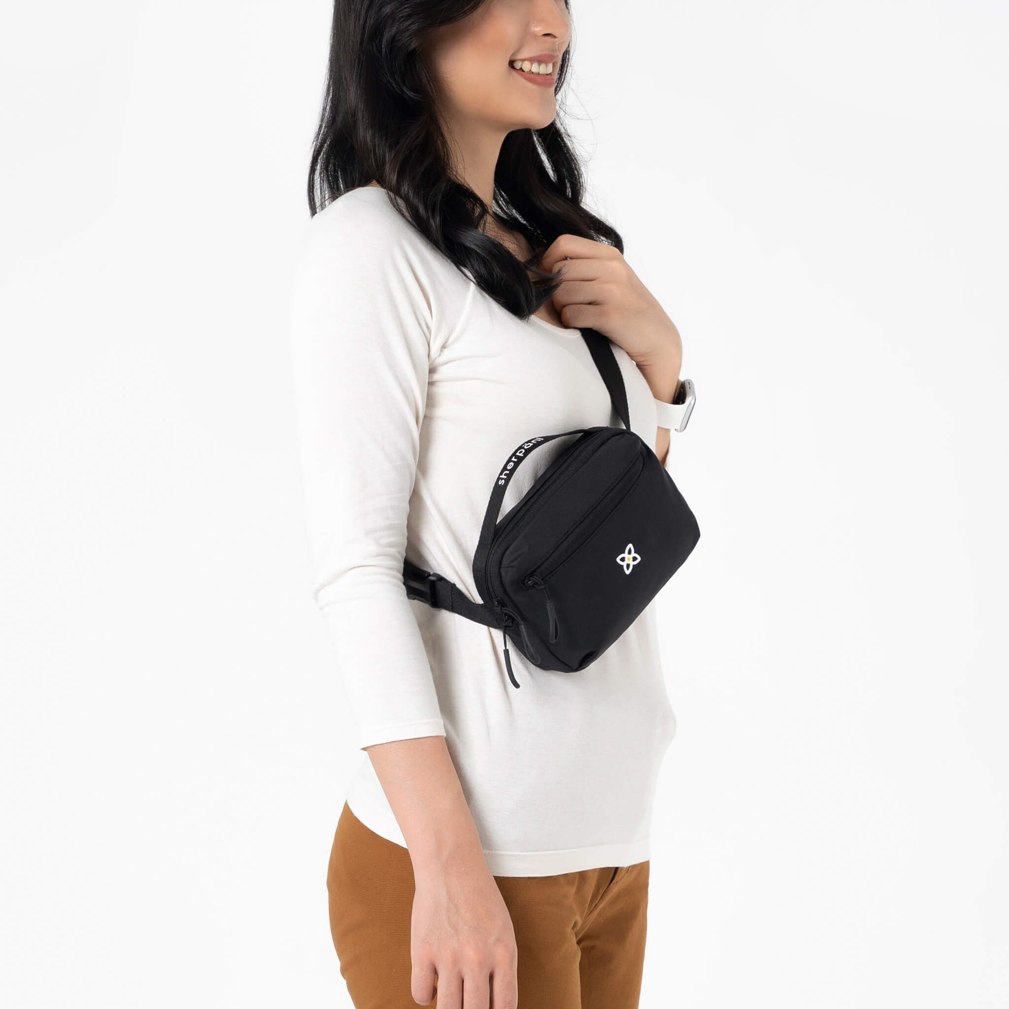 Close up view of a dark haired model smiling over her right shoulder. She is wearing a white tee shirt, brown pants and Sherpani&#39;s fanny pack, the Hyk in Raven, as a crossbody.