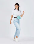 Full body view of a dark haired model smiling over her right shoulder. She is wearing a white tee shirt, faded jeans, white shoes and Sherpani's fanny pack, the Hyk in Seagreen, as a fanny pack.