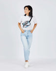 Full body view of a dark haired model facing the camera and smiling over her right shoulder. She is wearing a white tee shirt, faded jeans, white sneakers and Sherpani's fanny pack, the Hyk in Summer Camo, as a crossbody.