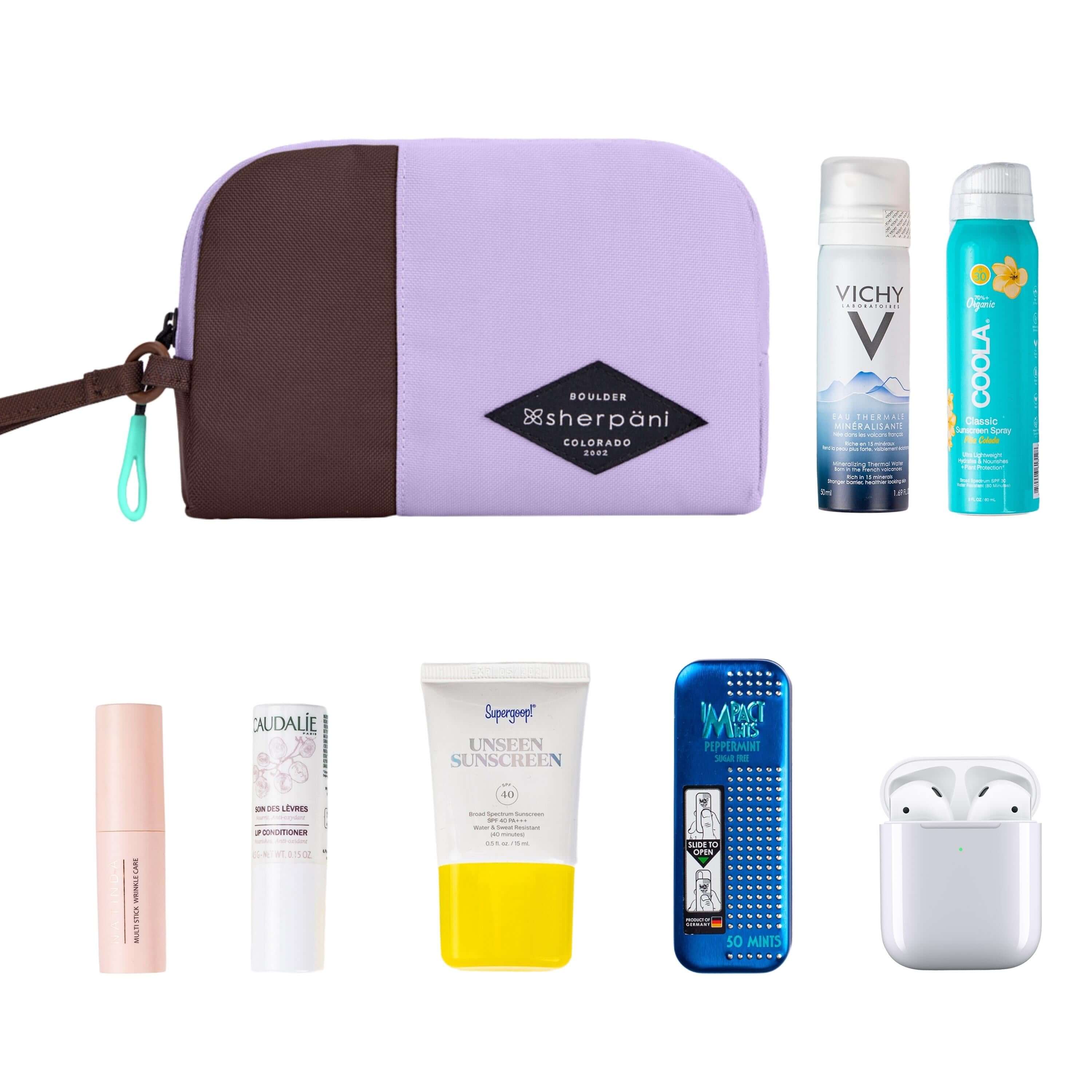 Top view of example items to fill the bag. Sherpani travel accessory, the Jolie in Lavender in medium size, lies in the upper left corner. It is surrounded by an assortment of items: beauty products, skincare products, sunscreen, mints and AirPods. 