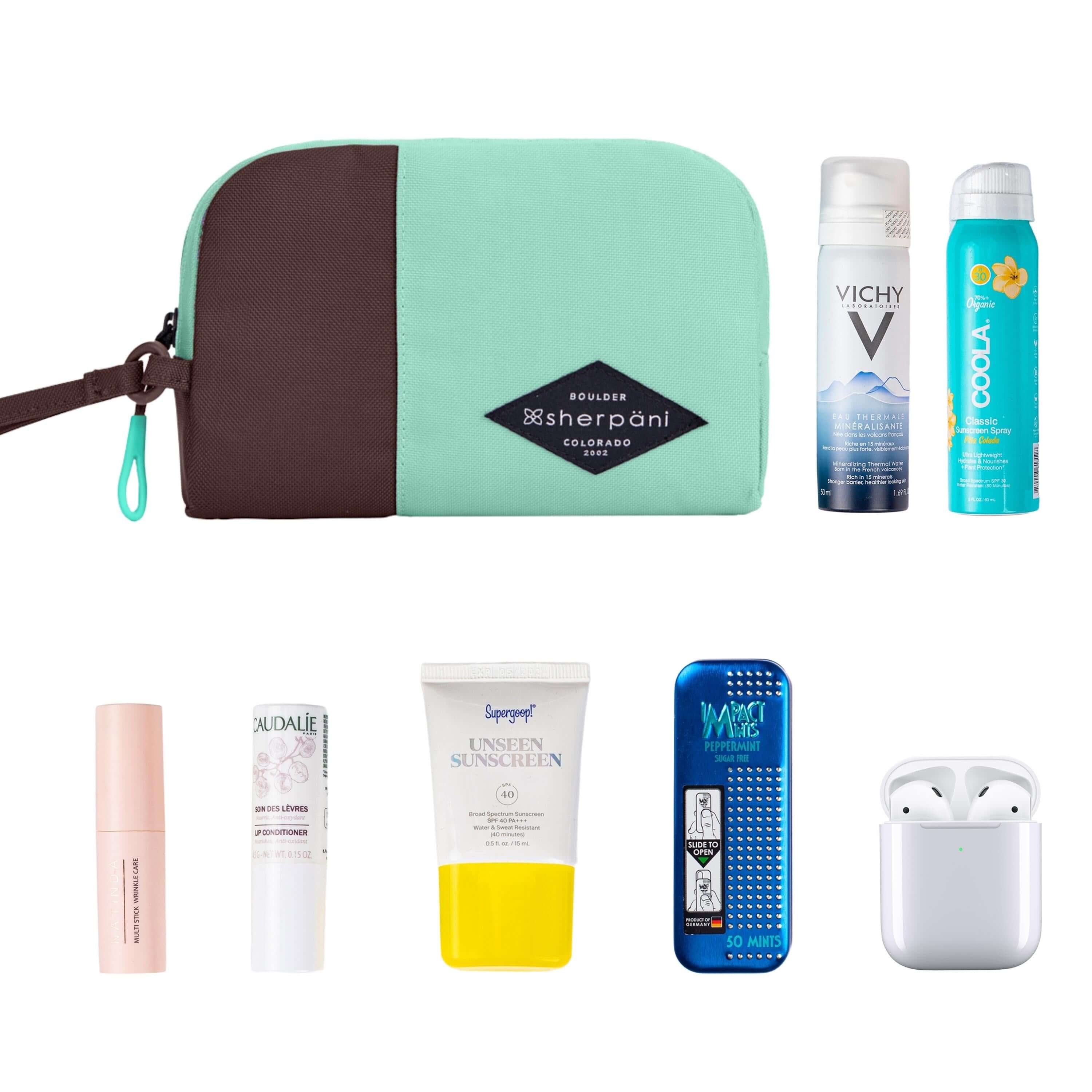 Top view of example items to fill the bag. Sherpani travel accessory, the Jolie in Seagreen in medium size, lies in the upper left corner. It is surrounded by an assortment of items: beauty products, skincare products, sunscreen, mints and AirPods.