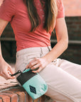 A brown haired woman sits outside on a brick ledge. She is wearing a salmon-colored top and tan pants. By her hip, she is unzipping Sherpani travel accessory the Jolie in Seagreen.