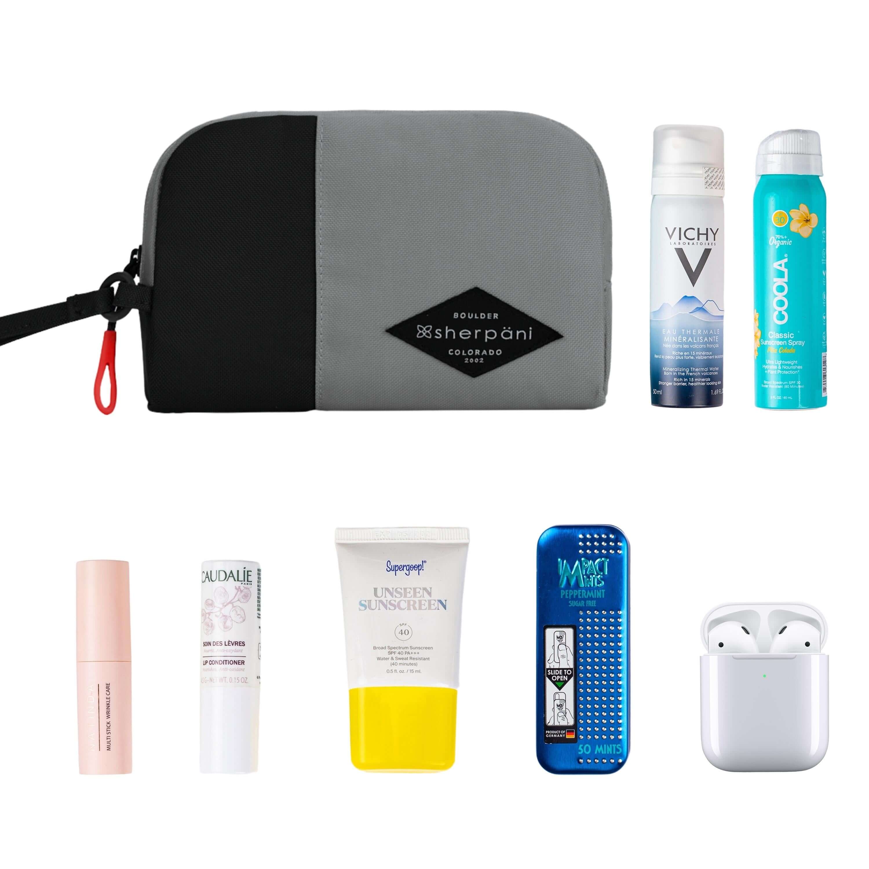 Top view of example items to fill the bag. Sherpani travel accessory, the Jolie in Stone in medium size, lies in the upper left corner. It is surrounded by an assortment of items: beauty products, skincare products, sunscreen, mints and AirPods.