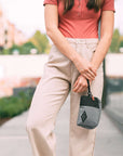 A brown haired woman stands outside on a path. She is wearing a salmon-colored top and tan pants. She carries Sherpani travel accessory, the Jolie in Stone, as a wristlet.
