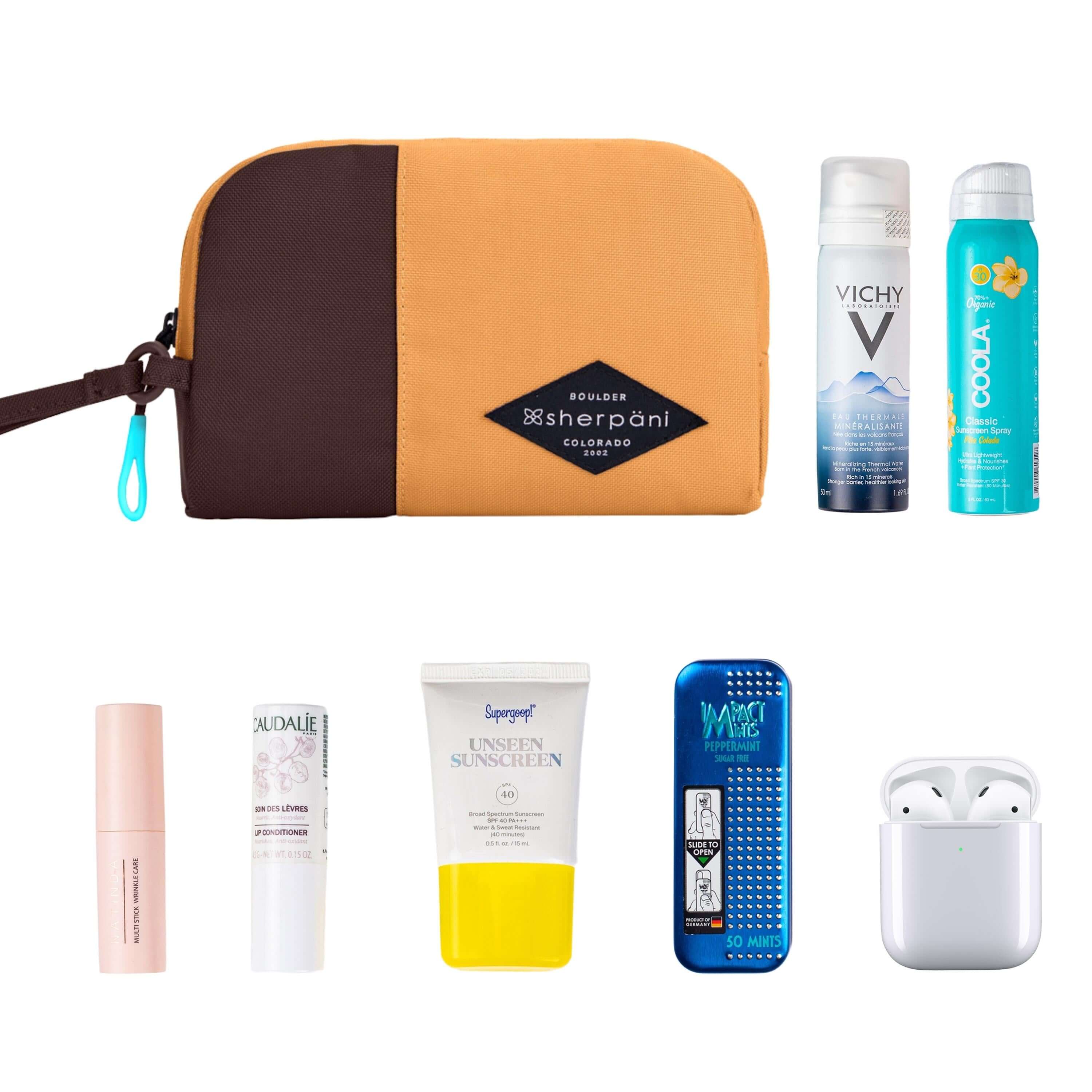 Top view of example items to fill the bag. Sherpani travel accessory, the Jolie in Sundial in medium size, lies in the upper left corner. It is surrounded by an assortment of items: beauty products, skincare products, sunscreen, mints and AirPods.