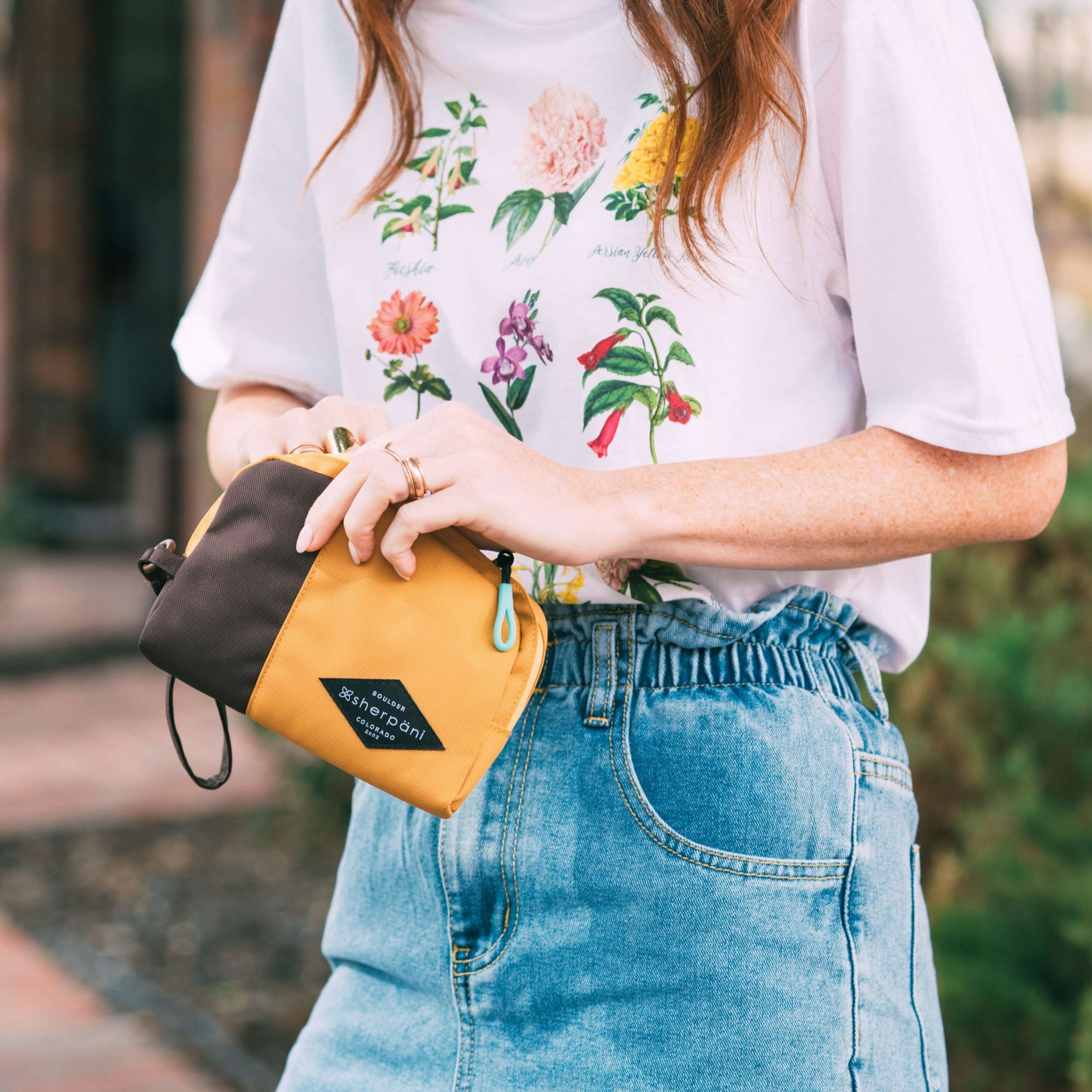 A red haired woman stands outside. She is wearing a white shirt with flowers and jeans. She is opening Sherpani travel accessory, the Jolie in Sundial.