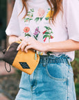 A red haired woman stands outside. She is wearing a white shirt with flowers and jeans. She is opening Sherpani travel accessory, the Jolie in Sundial.