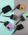 Top view of six Jolie travel pouches on a green and gray background. Clockwise form upper left: medium size Jolie in Raven, small size Jolie in Sundial, medium size Jolie in Seagreen, small size Jolie in Chromatic, medium size Jolie in Lavender, small size Jolie in Stone.