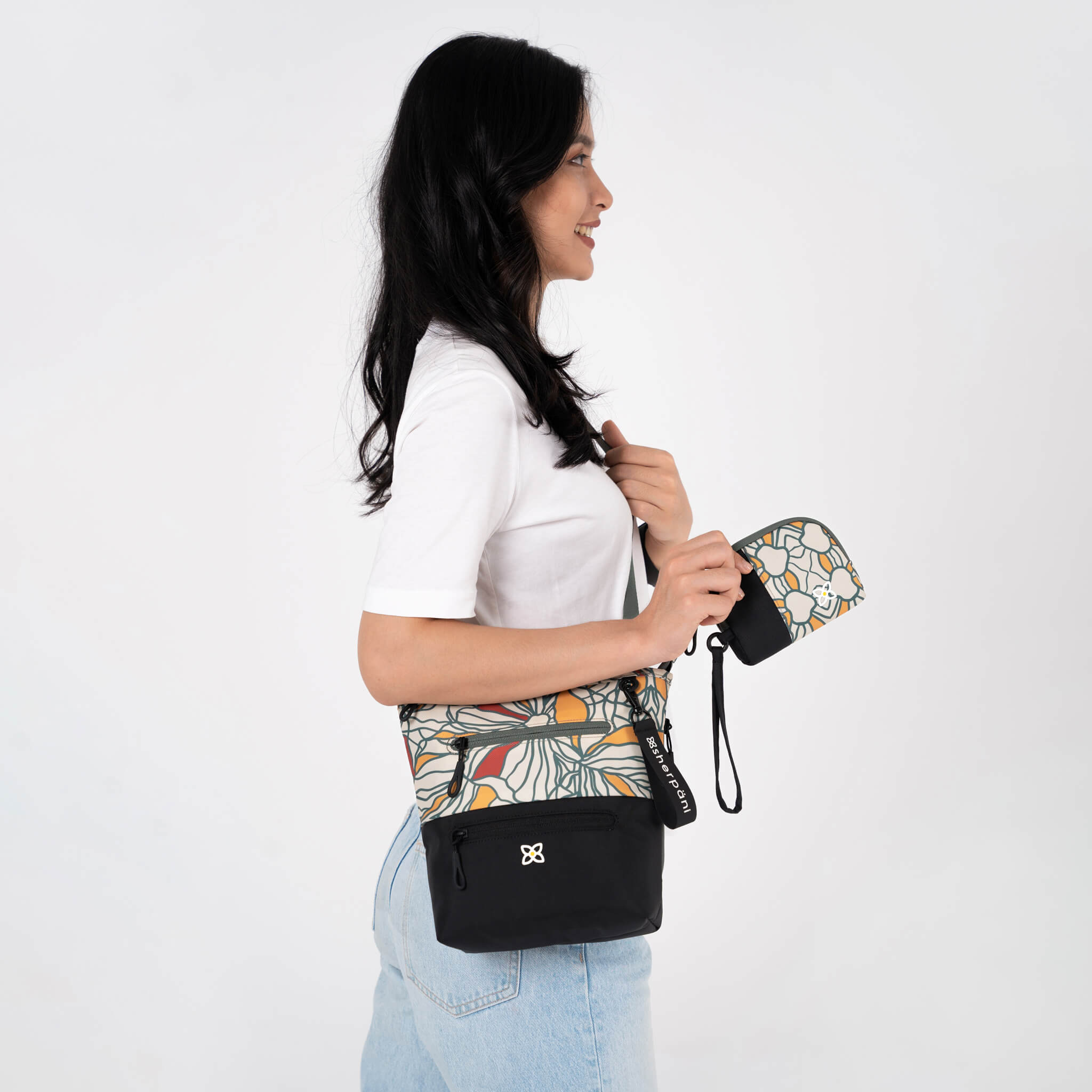A model is wearing Sherpani crossbody travel purse, the Sadie in Fiori. She is also holding Sherpani mini travel wristlet, the Jolie in Fiori. 