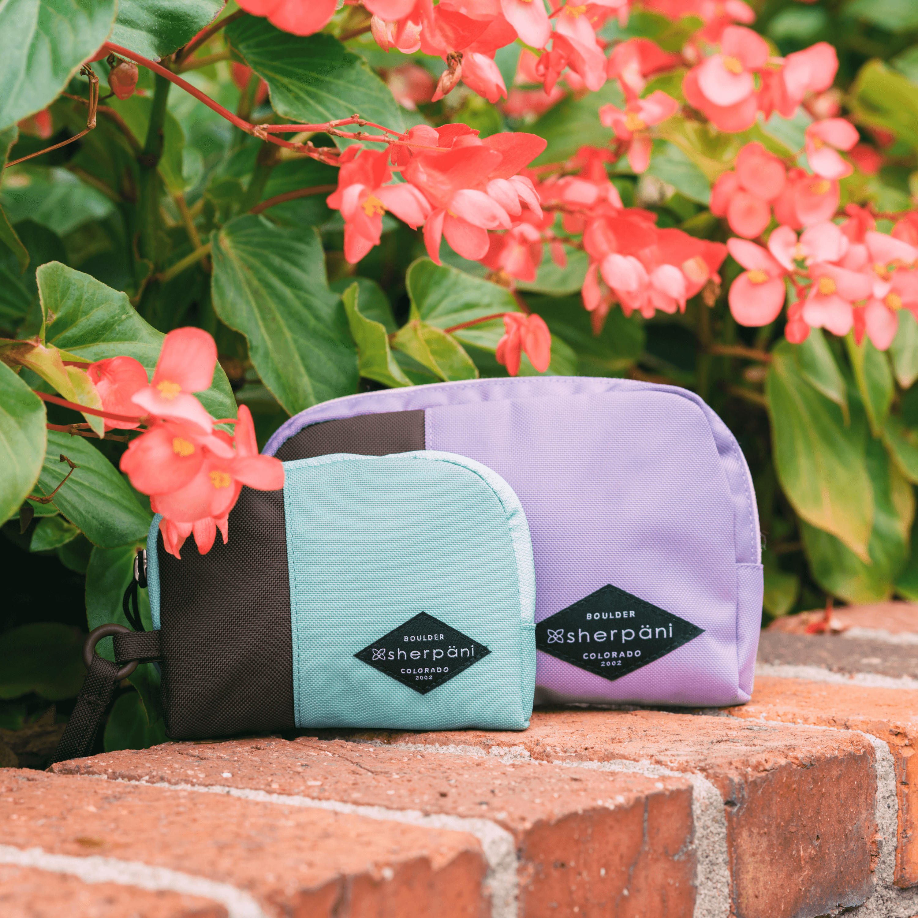 Two Sherpani travel accessories sit on a brick ledge outside under some flowers. A medium size Jolie in Lavender and small size Jolie in Seagreen.