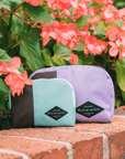 Two Sherpani travel accessories sit on a brick ledge outside under some flowers. A medium size Jolie in Lavender and small size Jolie in Seagreen.