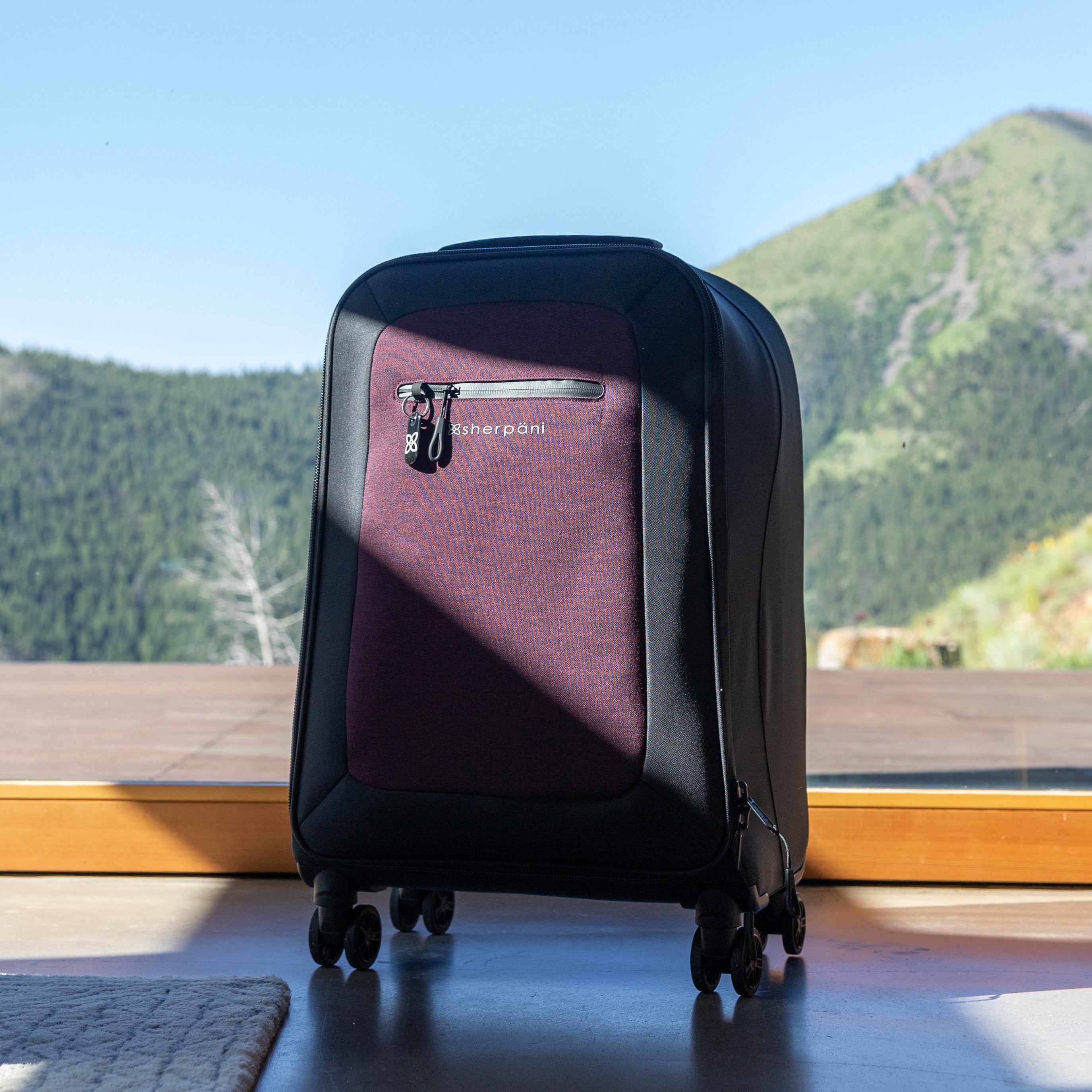 Sherpani soft-shell carry-on luggage, the Latitude in Merlot, sits in front of a window looking out over a scenic mountain view.