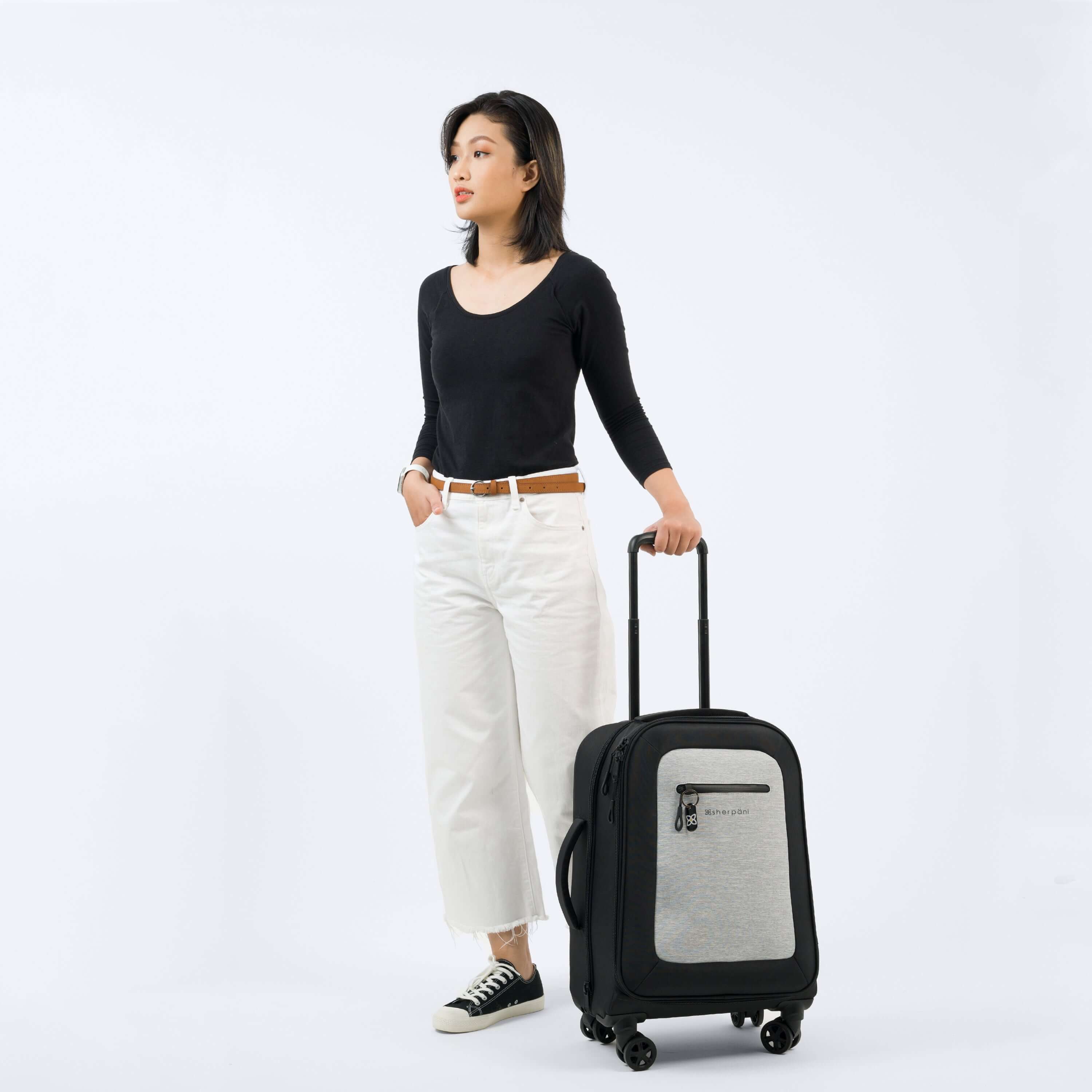 A dark haired woman stands at an angle to the camera. She is wearing a black top, white jeans and Converse. Her left hand holds the luggage handle of Sherpani&#39;s Anti-Theft luggage, the Latitude in Sterling, which stands next to her.