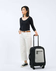 A dark haired woman stands at an angle to the camera. She is wearing a black top, white jeans and Converse. Her left hand holds the luggage handle of Sherpani's Anti-Theft luggage, the Latitude in Sterling, which stands next to her.
