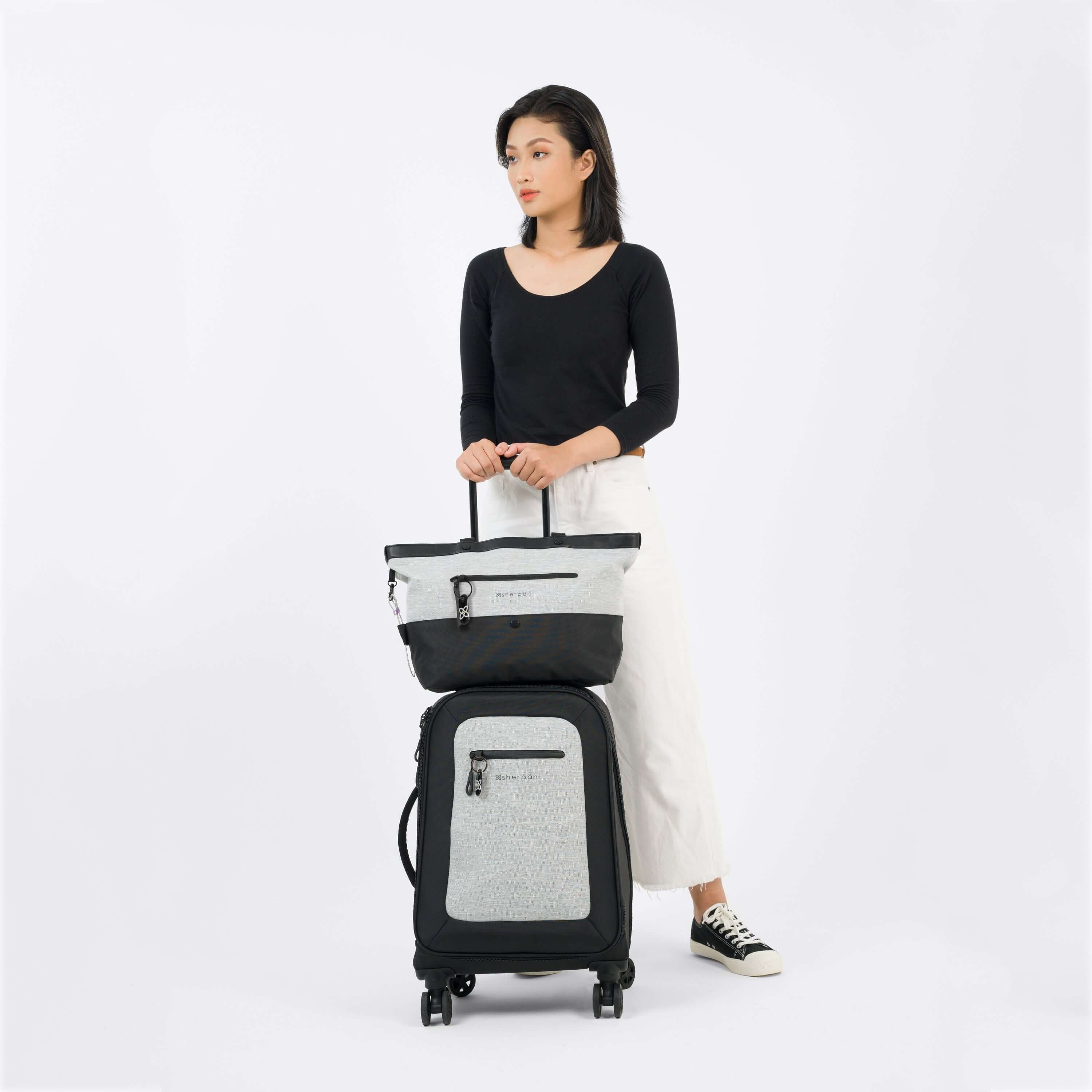 Full body view of a dark haired model wearing a black top, white jeans and Converse. She is standing behind Sherpani&#39;s Anti-Theft luggage, the Latitude in Sterling, and holding onto the retractable luggage handle. Sherpani&#39;s Anti-Theft tote, the Cali in Sterling, sits atop the suitcase, utilizing the luggage pass-through.