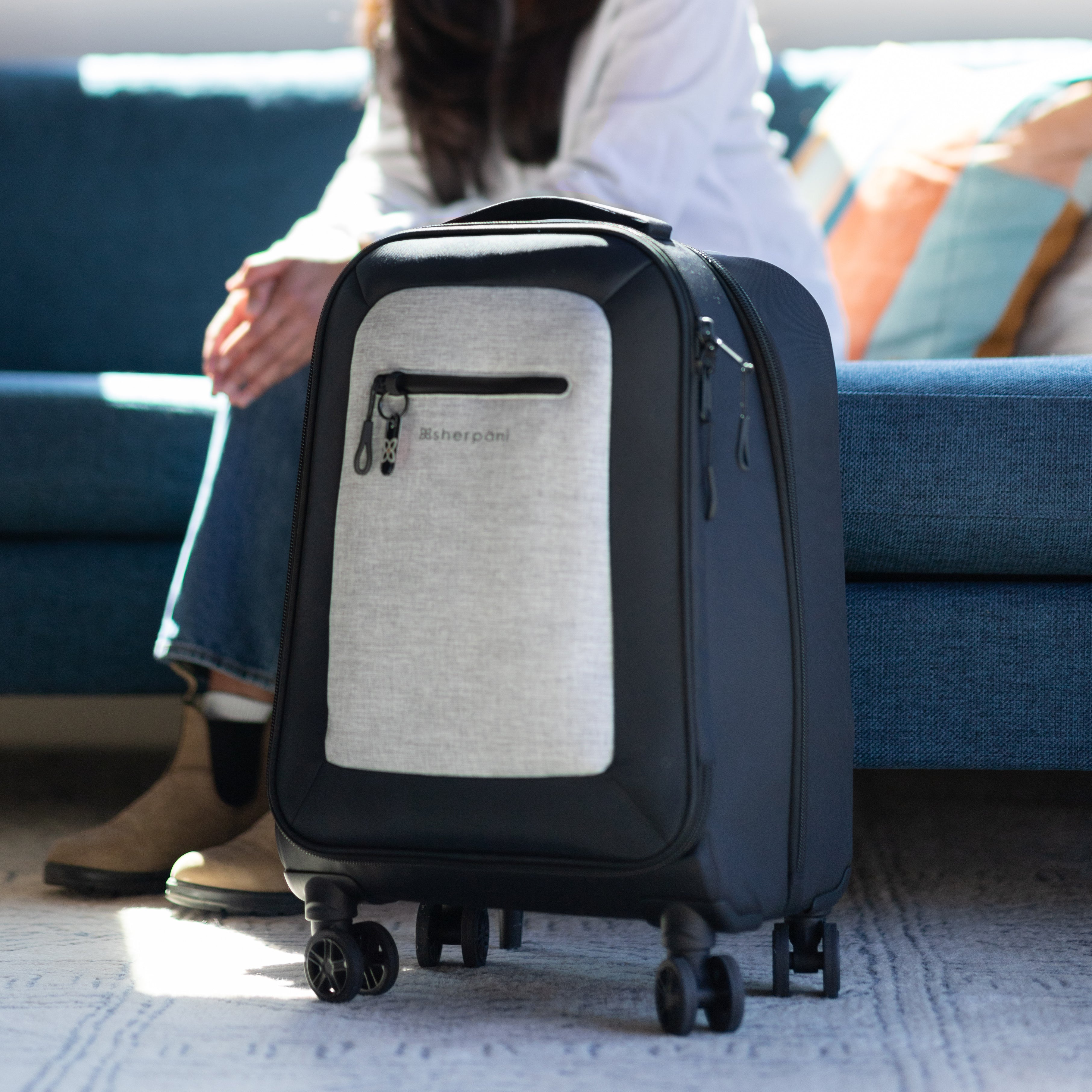 A woman sits on a couch behind Sherpani soft-shell carry-on luggage, the Latitude in Sterling.
