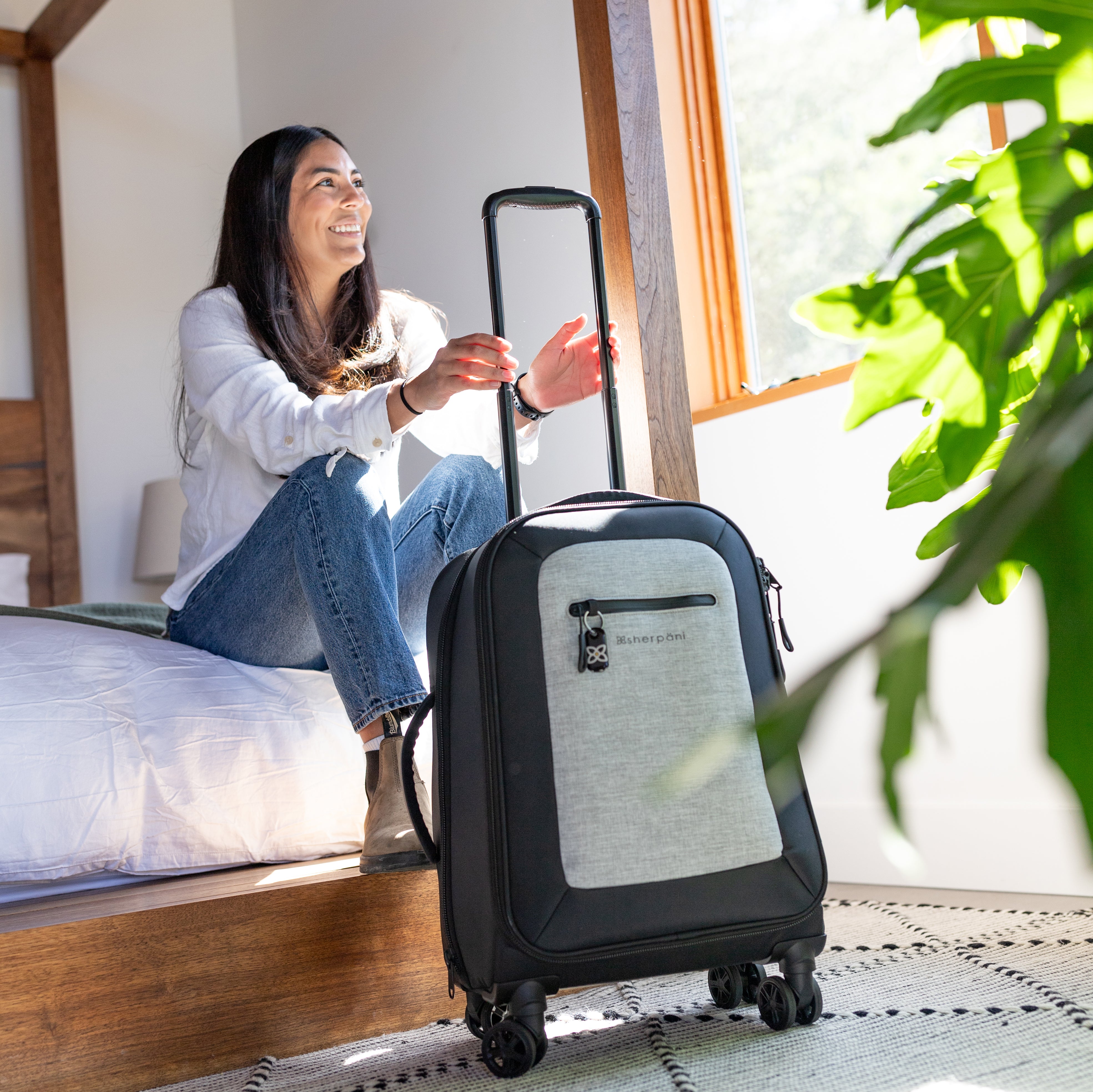 A dark-haired woman sits in on the bed of her hotel room in front of Sherpani soft-shell carry-on luggage, the Latitude in Sterling. 
