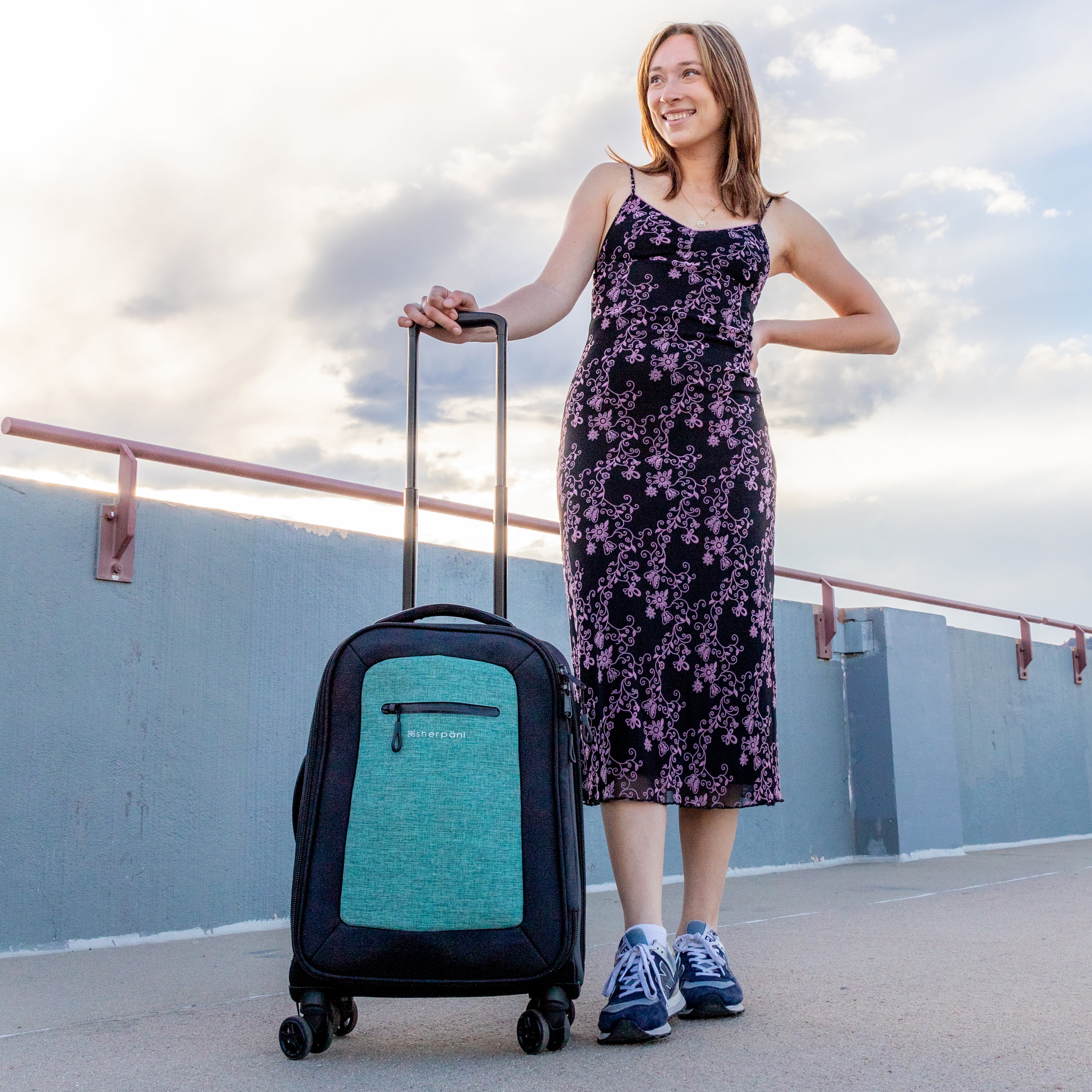 A woman stands in a parking lot. She is facing the camera and wearing a floral dress. By her side is Sherpani soft-shell carry-on luggage, the Latitude in Teal. 
