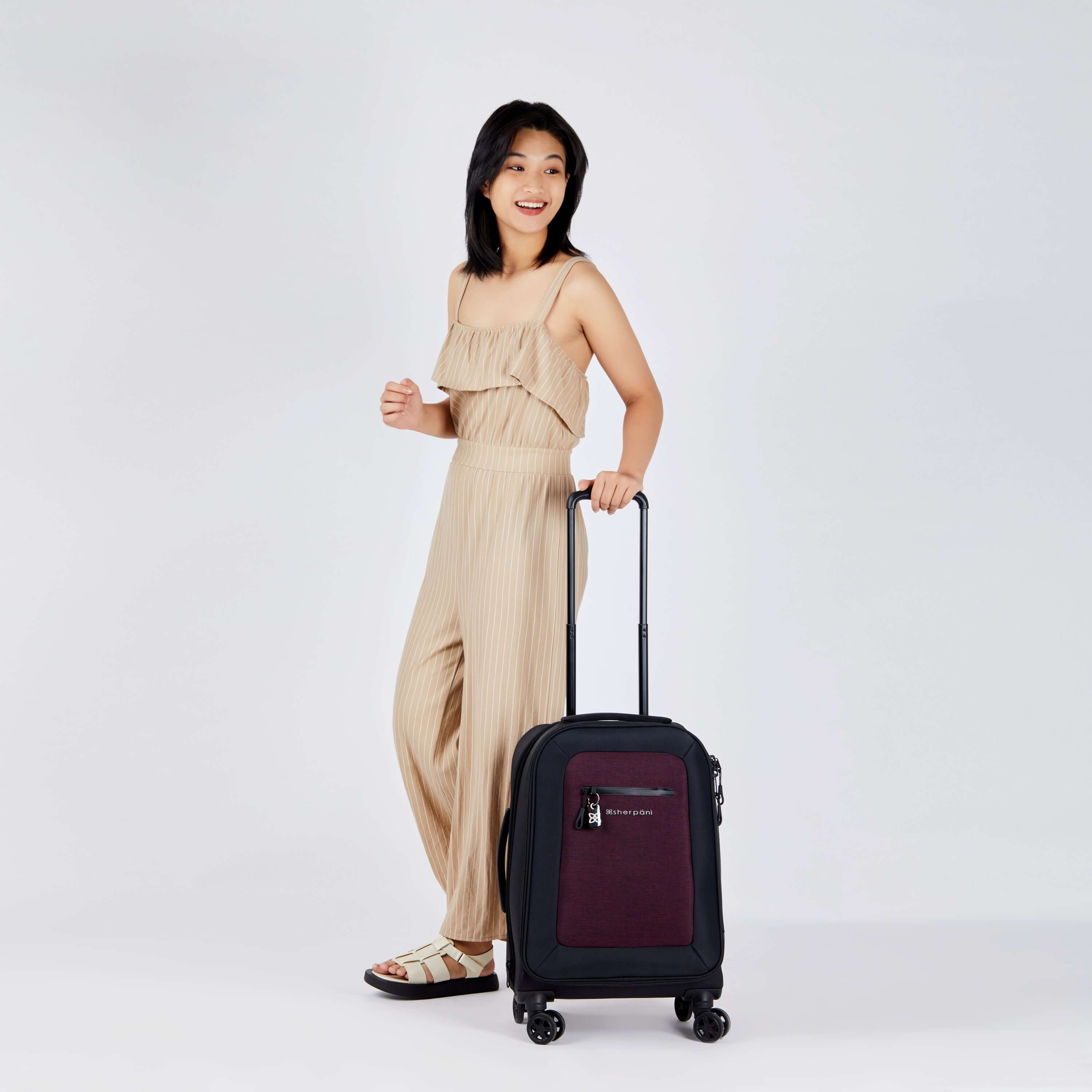 Full bodied view of a dark haired model facing to the side and smiling over her left shoulder. She is wearing a tan jumpsuit and sandals. Her left hand is holding the retractable luggage handle of Sherpani&#39;s Anti-Theft suitcase, the Latitude in Merlot.