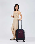 Full bodied view of a dark haired model facing to the side and smiling over her left shoulder. She is wearing a tan jumpsuit and sandals. Her left hand is holding the retractable luggage handle of Sherpani's Anti-Theft suitcase, the Latitude in Merlot.