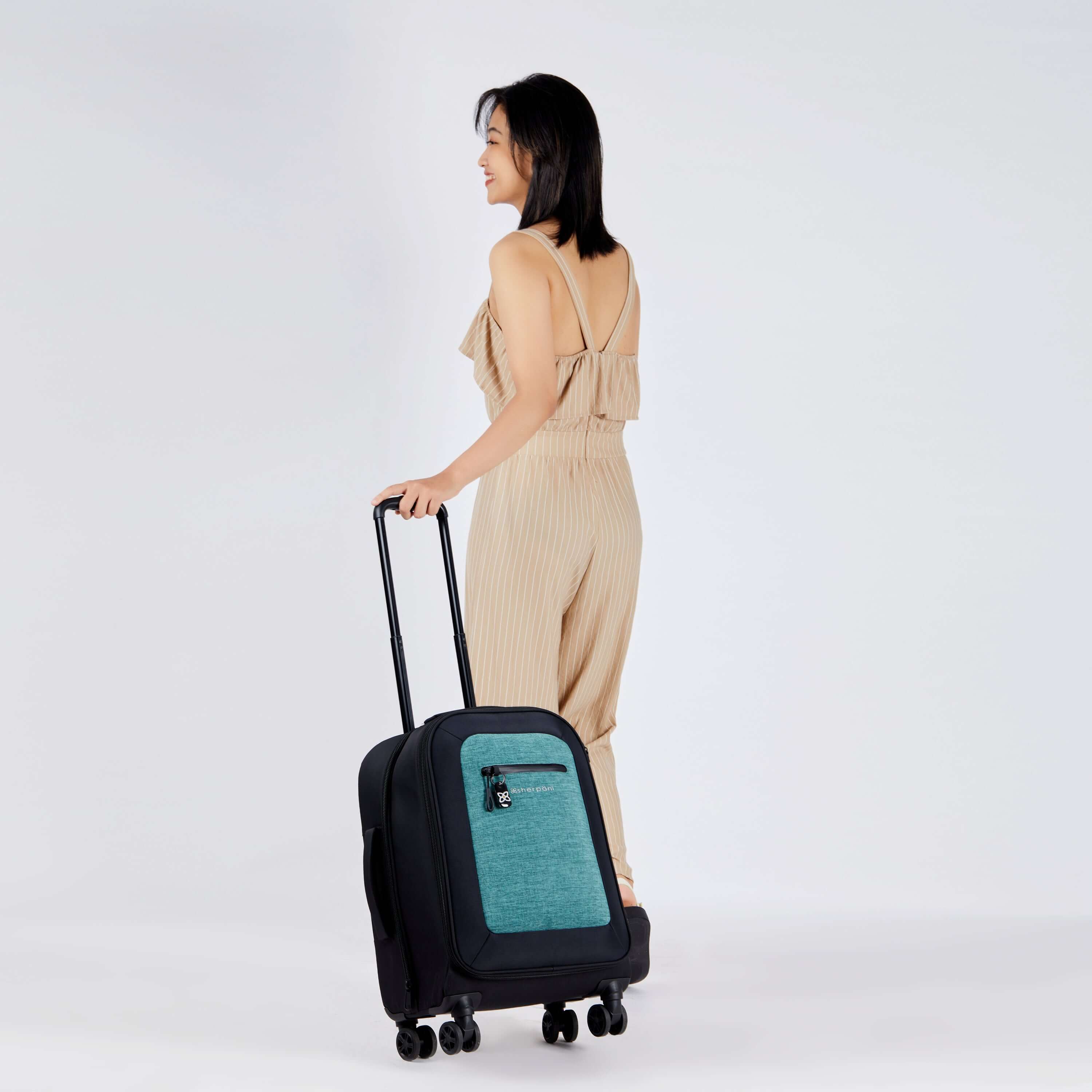 Full bodied view of a dark haired model facing away from the camera. She is wearing a tan jumpsuit. Sherpani's Anti-Theft luggage, the Latitude in Teal, rolls along beside her. 