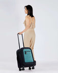 Full bodied view of a dark haired model facing away from the camera. She is wearing a tan jumpsuit. Sherpani's Anti-Theft luggage, the Latitude in Teal, rolls along beside her.