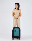 Full bodied view of dark haired model facing the camera and smiling over her right shoulder. She is wearing a tan jumpsuit. In front of her stands Sherpani's Anti-Theft luggage, the Latitude in Teal, which she is holding by the handle.