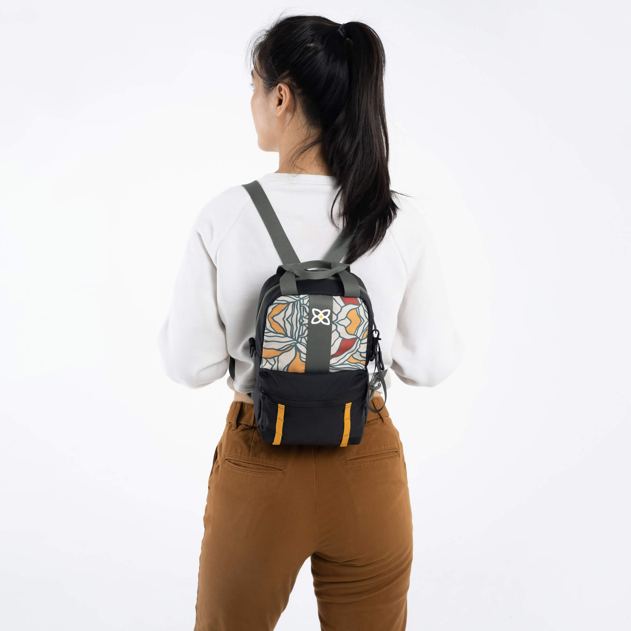 A model facing to the back. She is wearing a white top, brown pants and the Logan mini backpack in Fiori.
