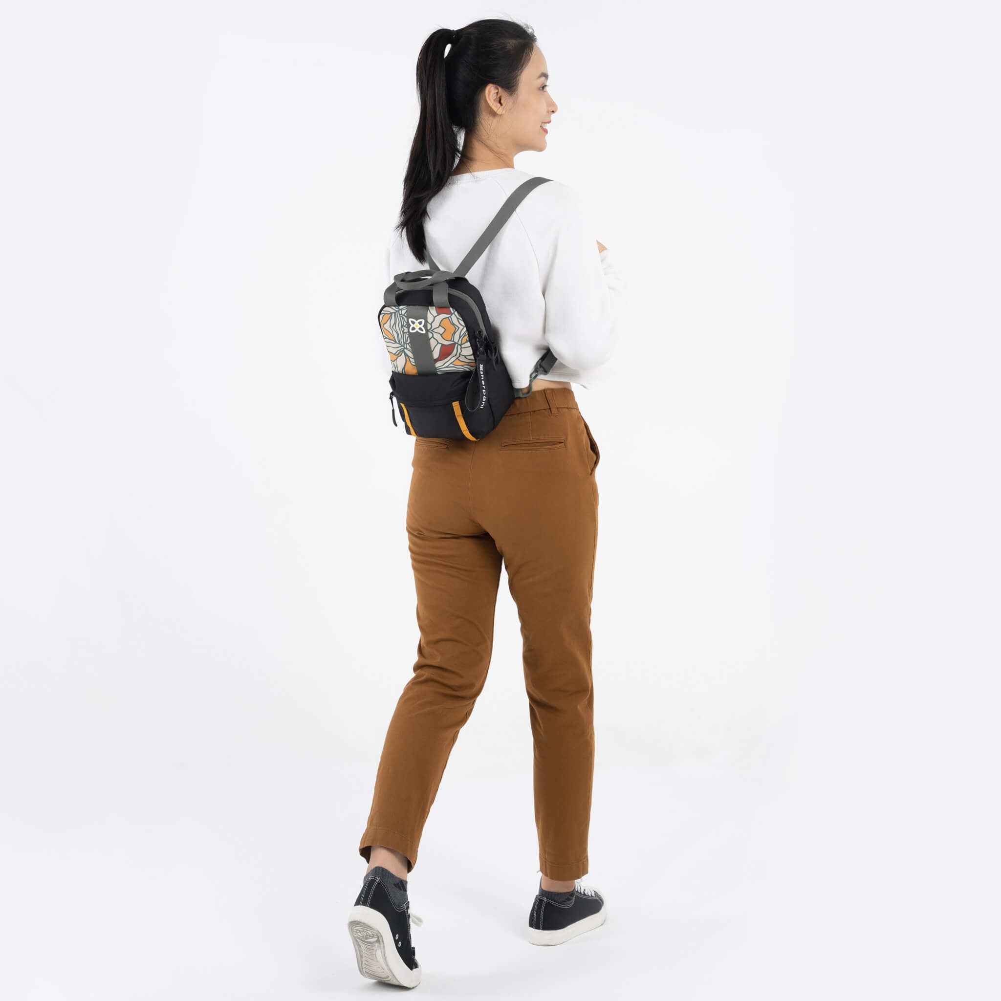 A model facing to the back. She is wearing a white top, brown pants and the Logan mini backpack in Fiori. 
