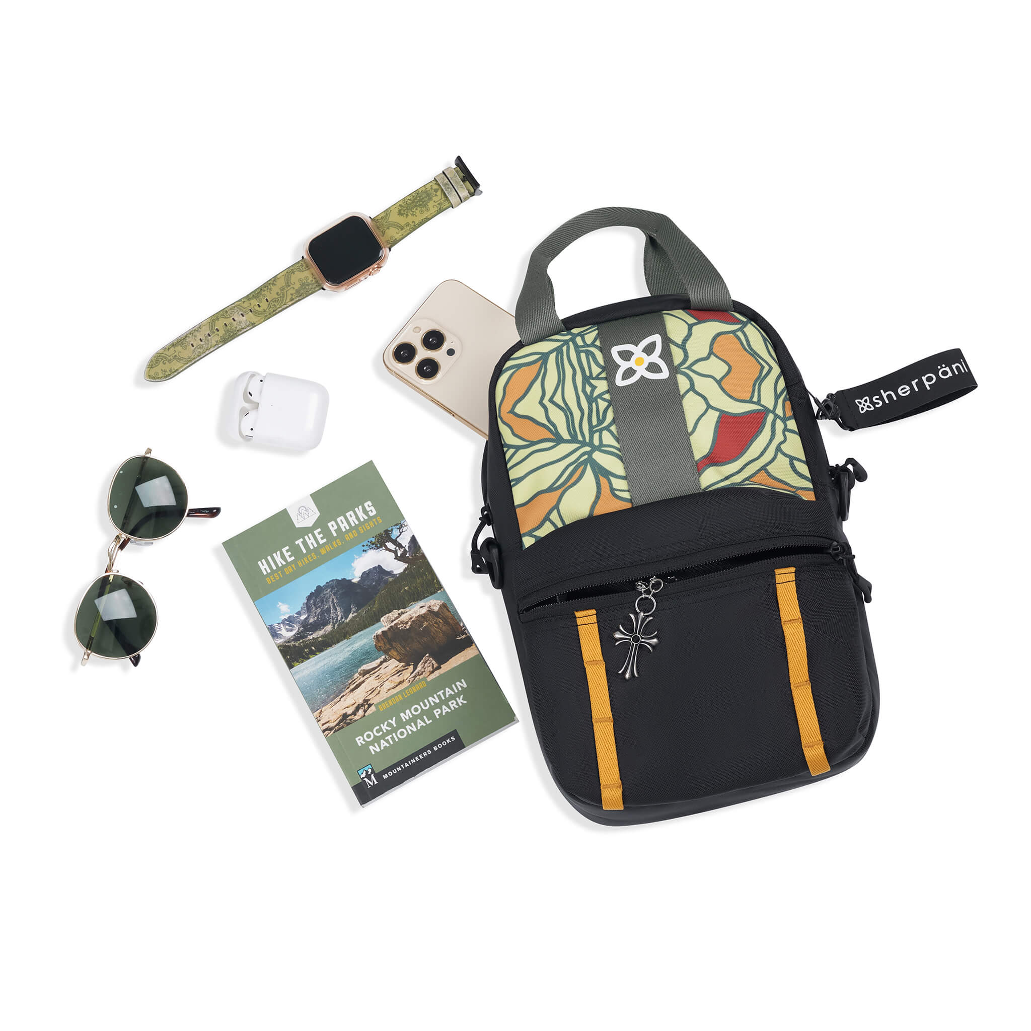 Top view of example items to fill the bag. Sherpani Logan mini backpack in Fiori lies next to the following items: smart watch, headphones, sunglasses, hiking guide and phone. 