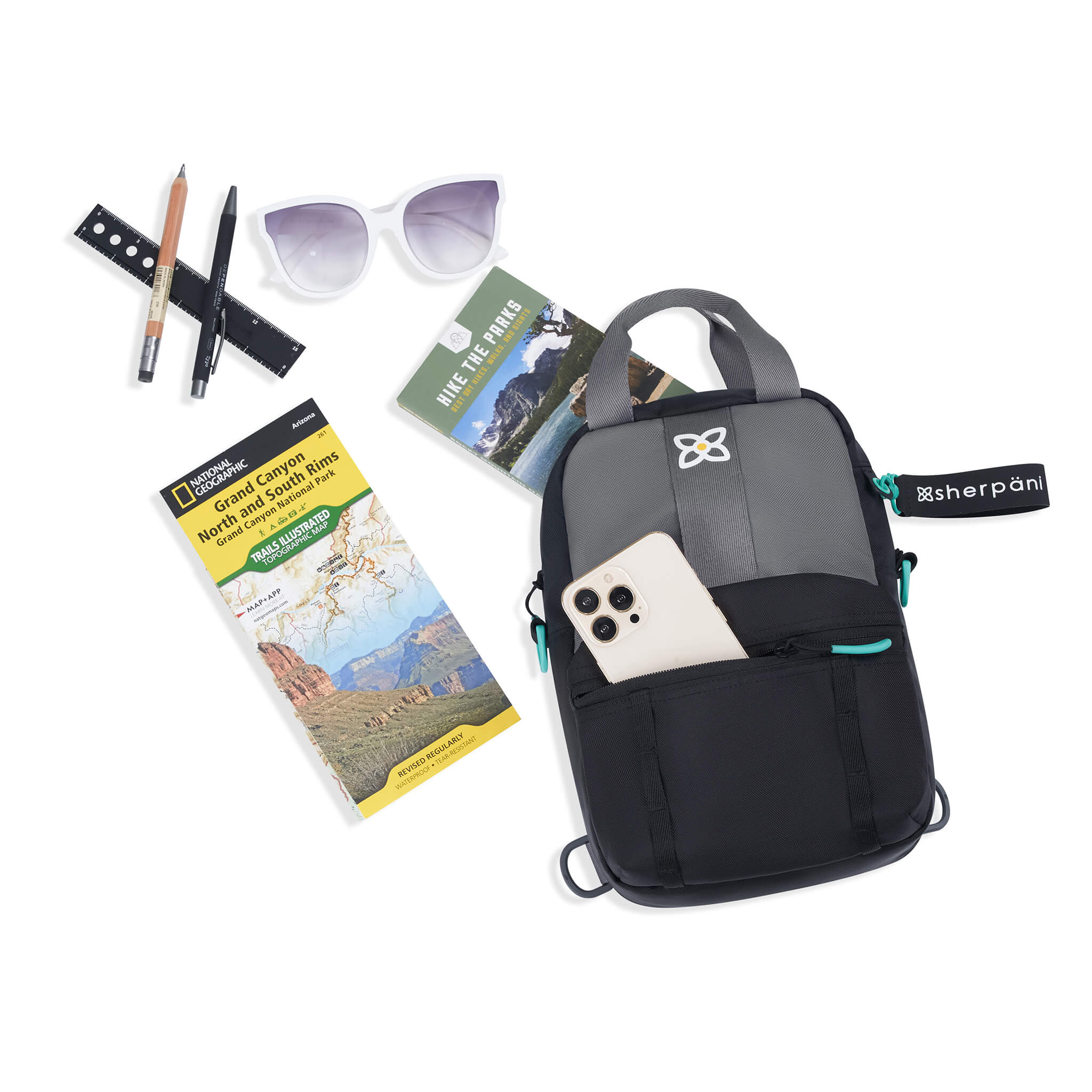  Top view of example items to fill the bag. Sherpani Logan mini backpack in Moonstone lies next to the following items: pencils, ruler, sunglasses, hiking guide, park map and phone.
