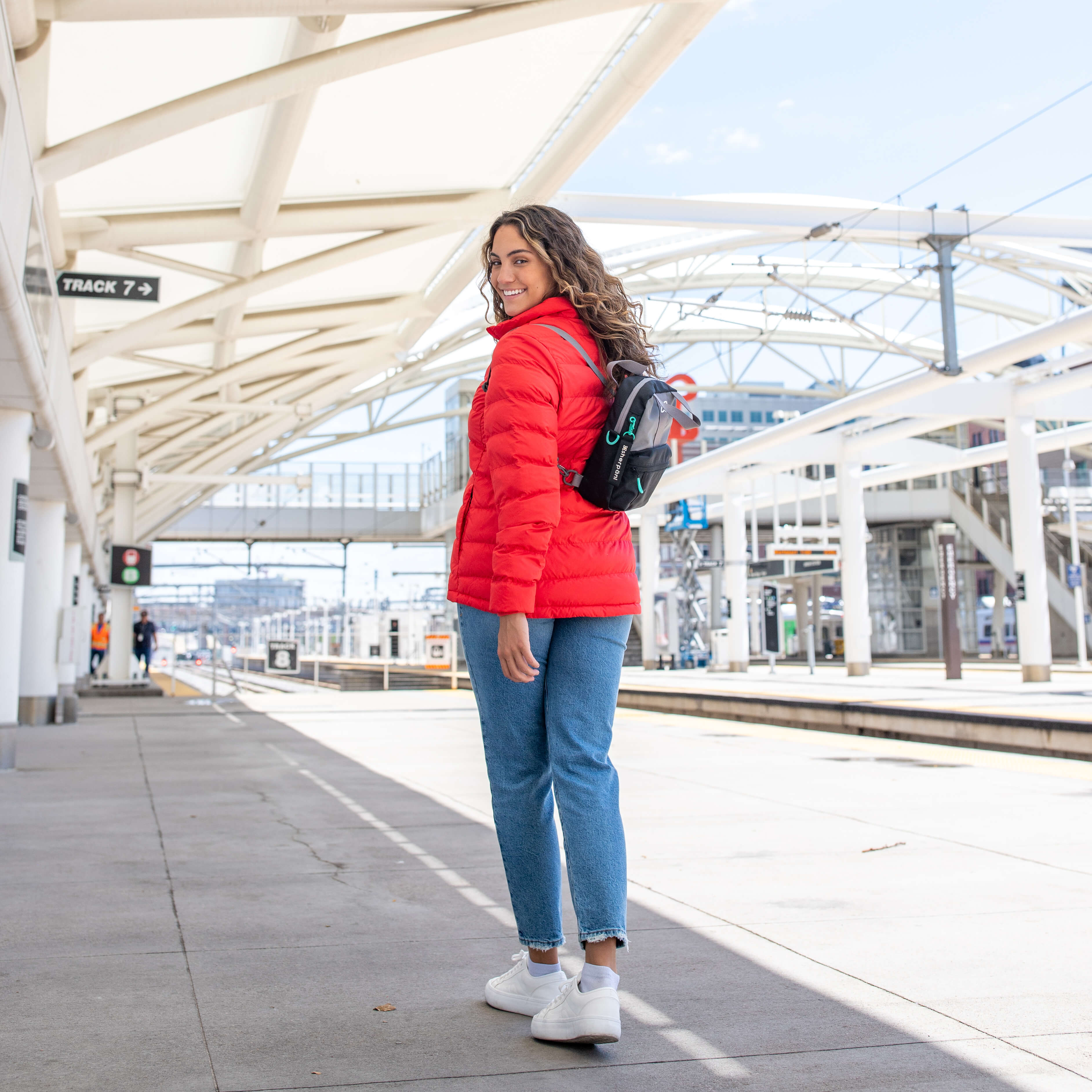 A traveling woman stands in a train station. She is wearing Sherpani small backpack for women, the Logan in Moonstone. 